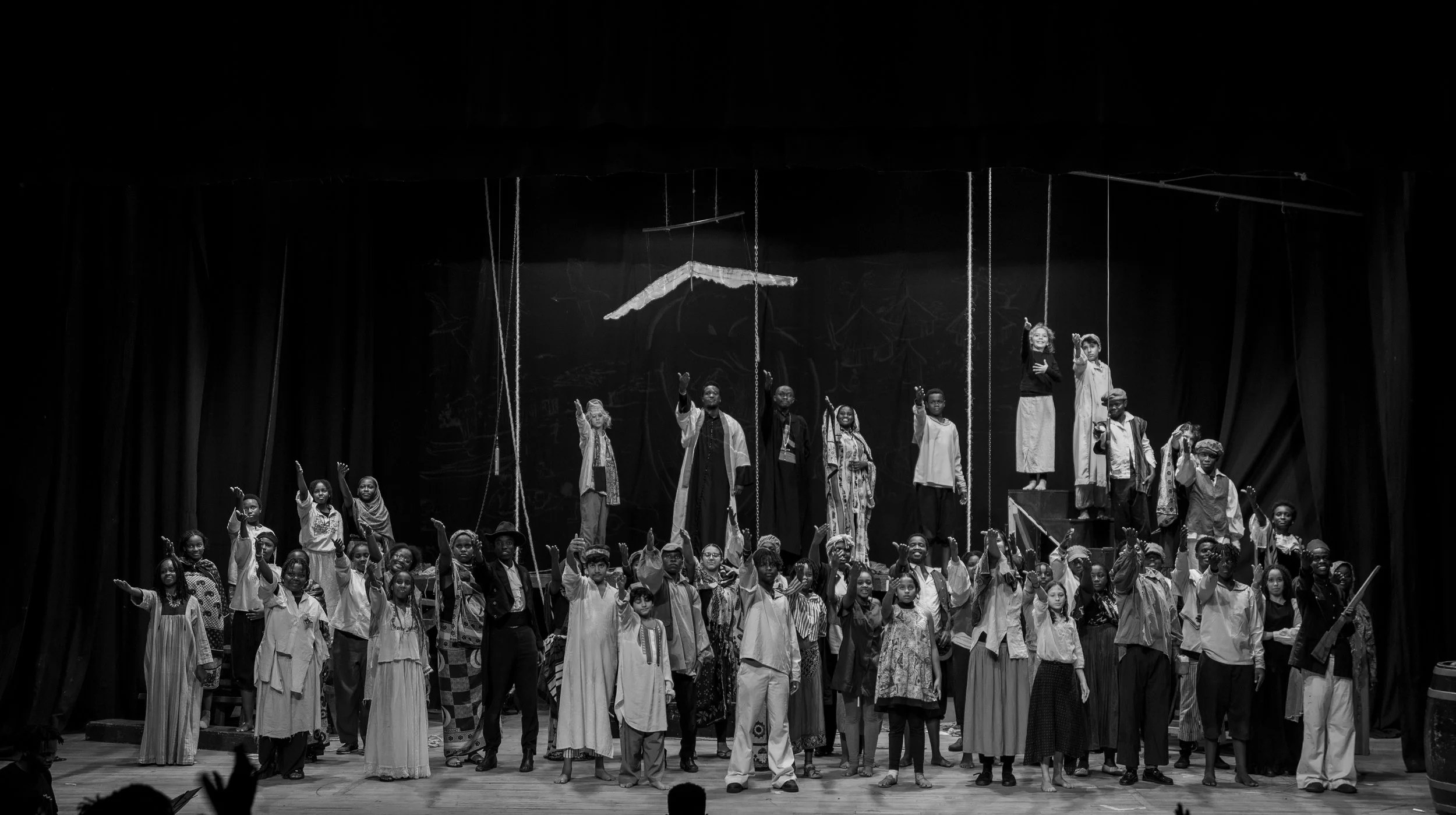 Group of children and adults on stage during a theatrical performance, many raising their hands, some wearing costumes, with a backdrop depicting a dark scene and a figure of a lion.