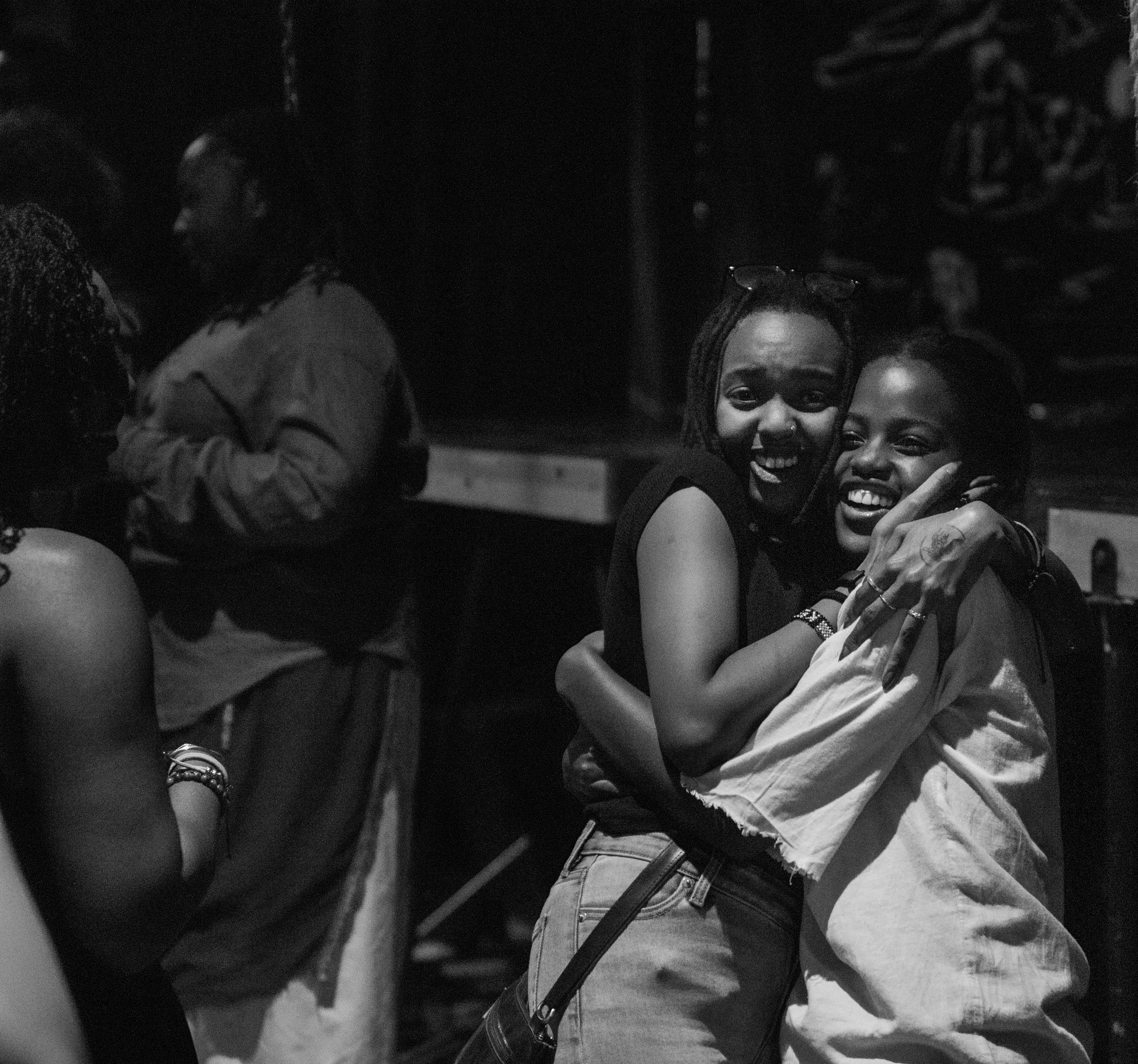 Two women with dark skin smiling and hugging each other, with other people in the background, in a dark setting.