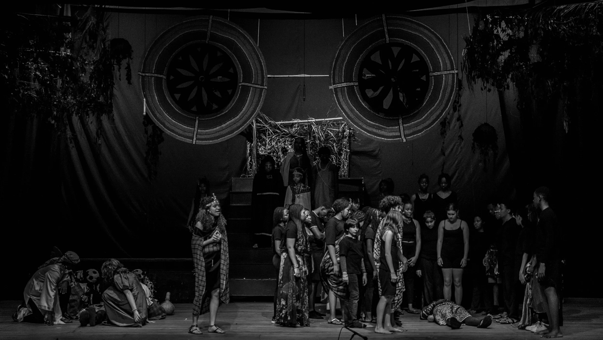 A theatrical performance on stage with a large group of people, including children and adults, some in costumes, in front of a dark backdrop with circular decorations.