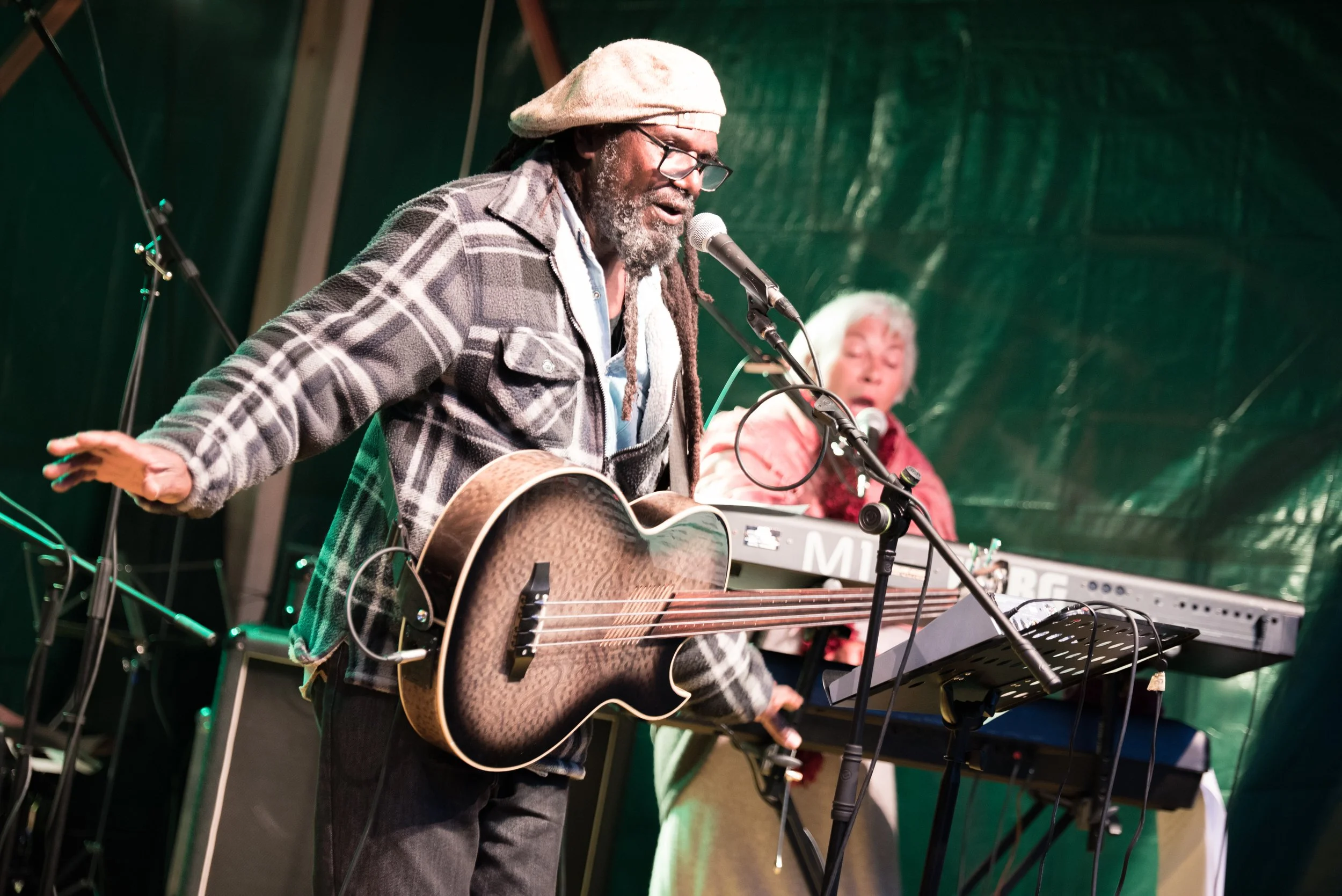 Deux musiciens jouent lors d'un concert, l'un avec une guitare acoustique et l'autre derrière un clavier, sous un abri vert.