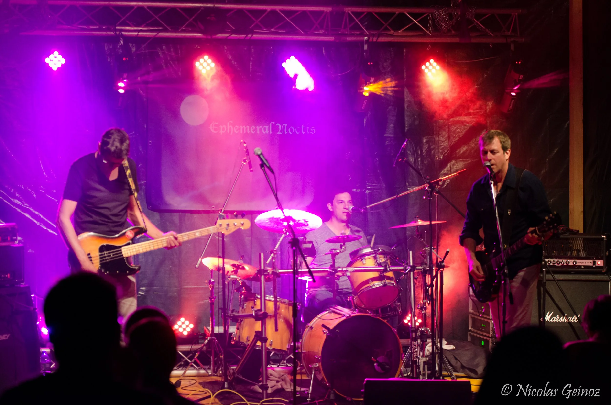 Un groupe de trois musiciens en concert, éclairage vibrant violet et rouge, scène avec instruments de musique, publique en silhouette.