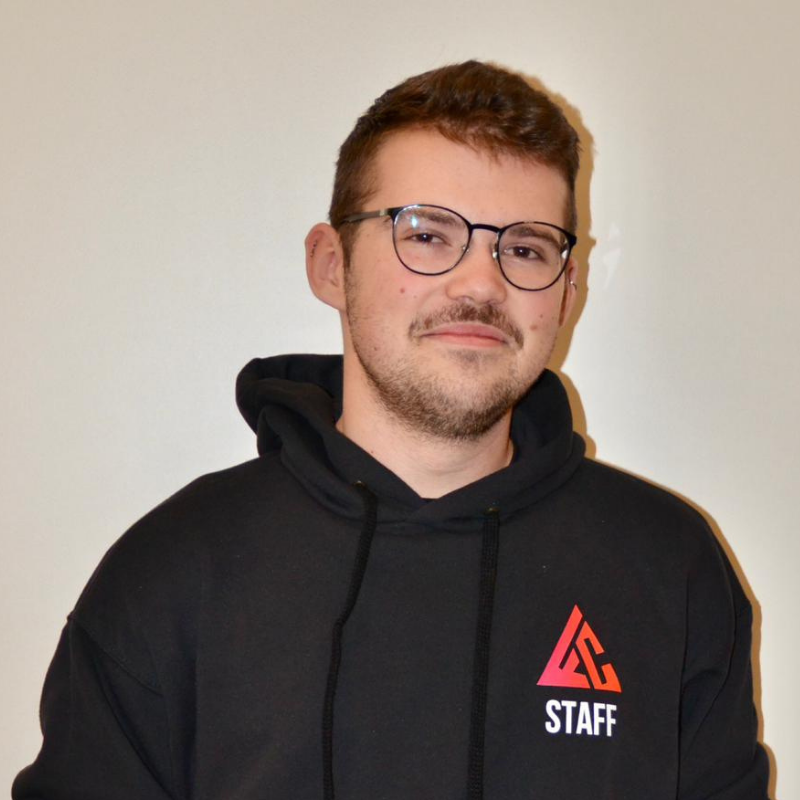 Un jeune homme portant un sweat à capuche noir avec un logo rouge et blanc, portant des lunettes, souriant légèrement, devant un mur blanc.