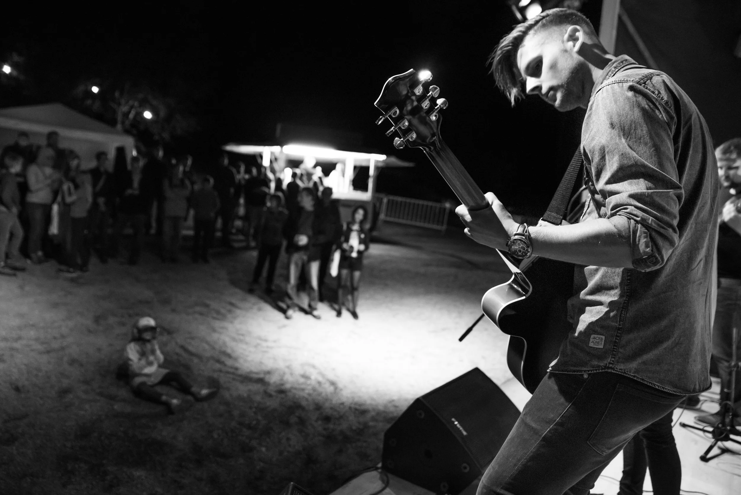 Un musicien joue de la guitare électrique lors d'un concert en plein air de nuit, avec un public devant lui. L'image est en noir et blanc.