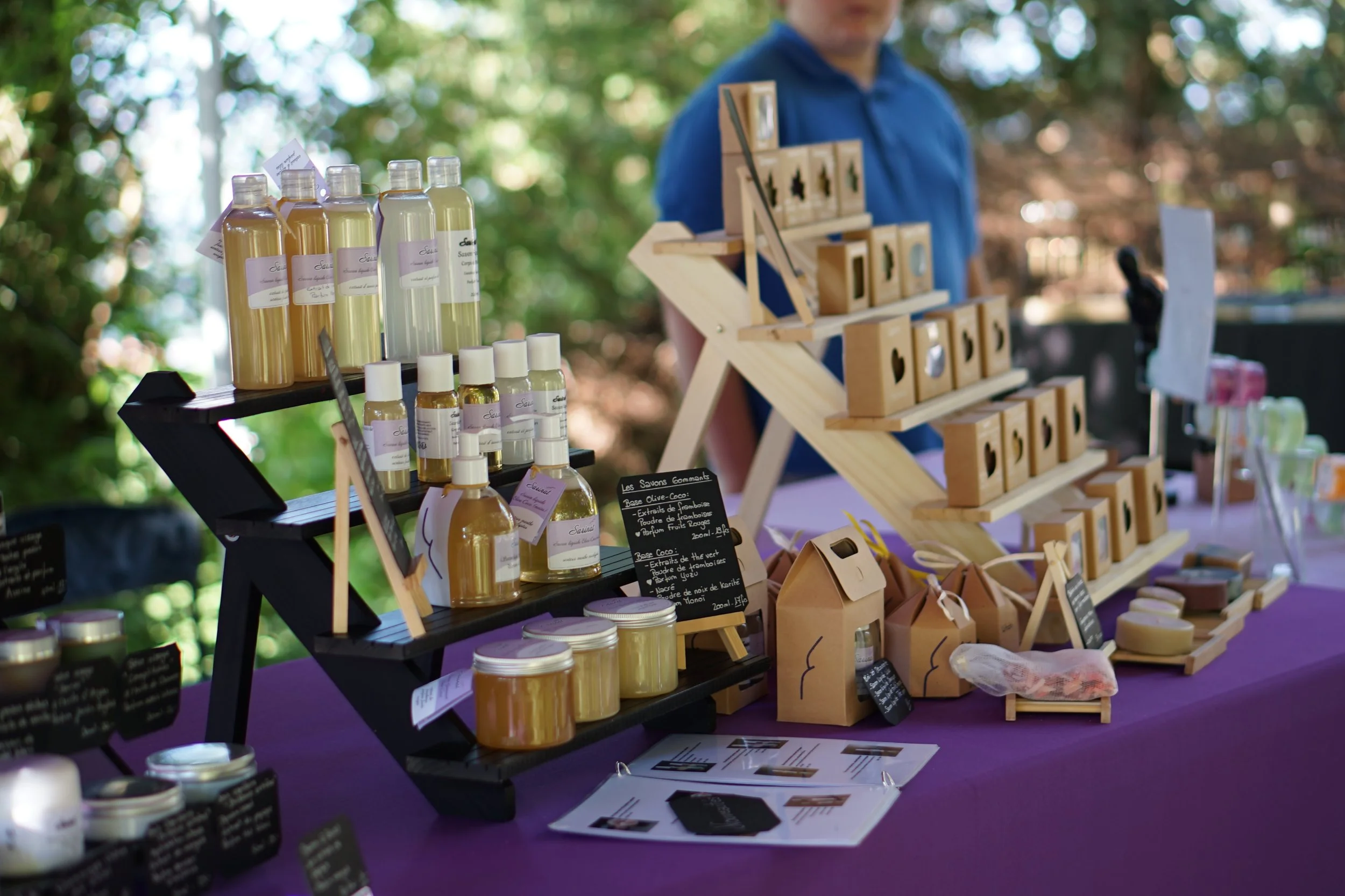 Table d'exposition avec des savons artisanaux, bouteilles de lotion, et des emballages en papier, lors d'un marché en plein air.