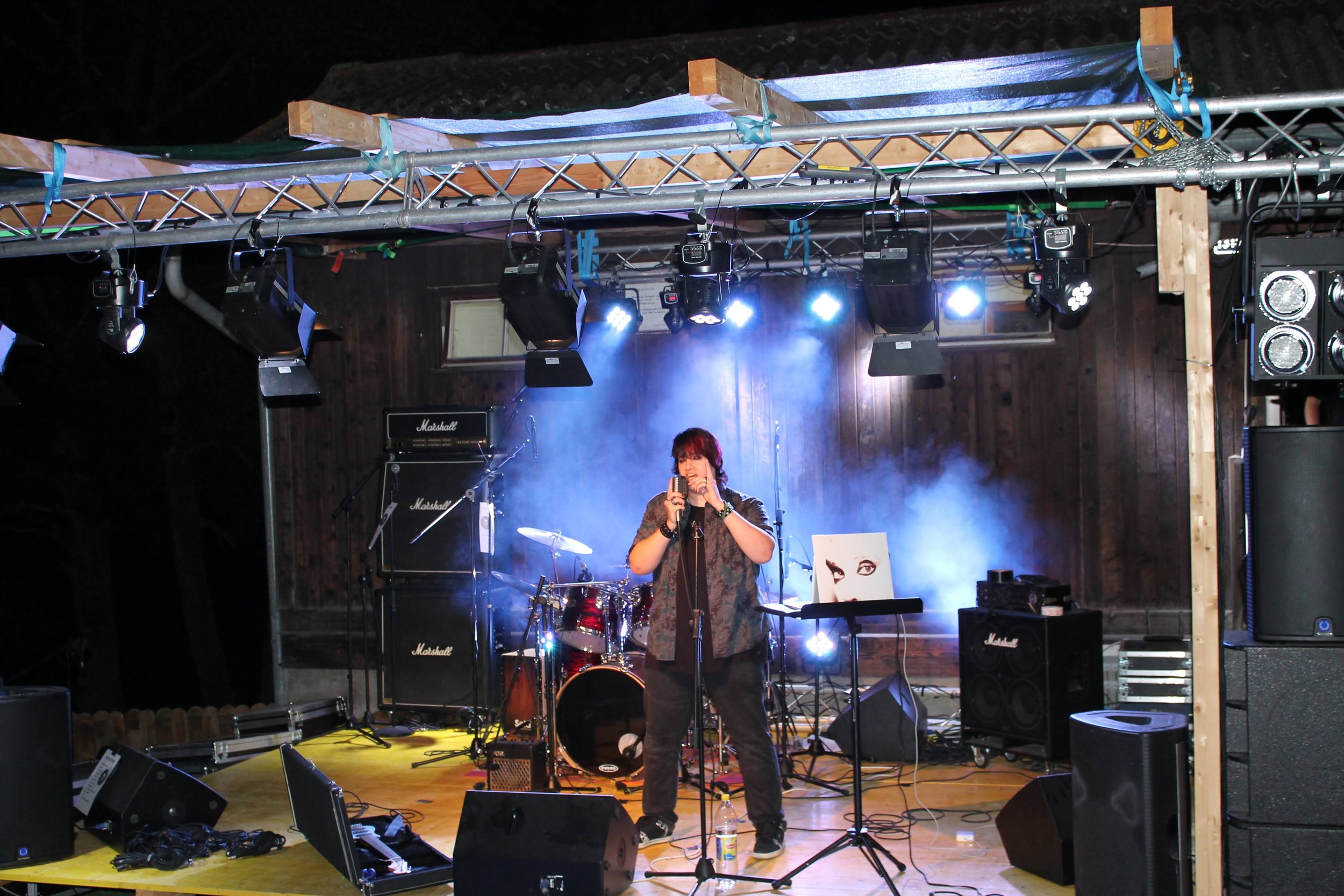 Une scène de concert avec un chanteur au centre, entouré d'instruments de musique, sous un éclairage de scène bleu et blanc, dans une ambiance sombre avec un fond en bois.