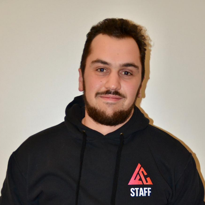 Un homme souriant avec une barbe, portant un sweat à capuche noir avec un logo 'STAFF' et un symbole coloré, posé devant un mur blanc.
