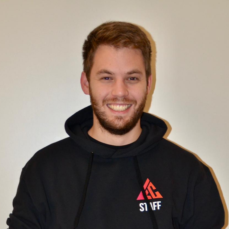 Un homme souriant portant un sweat à capuche noir avec un logo rouge et blanc, debout contre un mur clair.