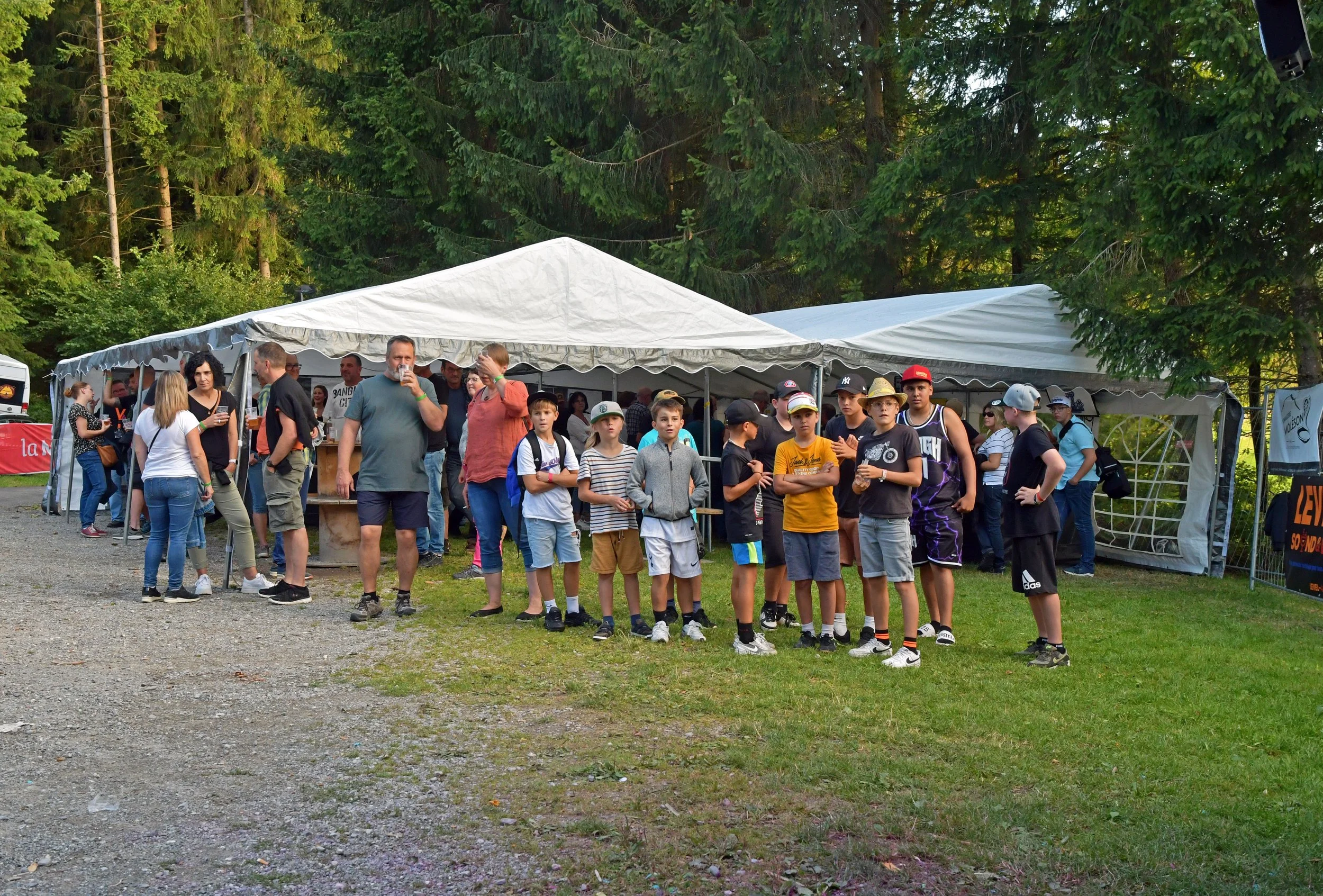 Groupe d'enfants et d'adultes en file devant un stand ou une tente lors d'un événement en plein air dans un espace vert avec des arbres