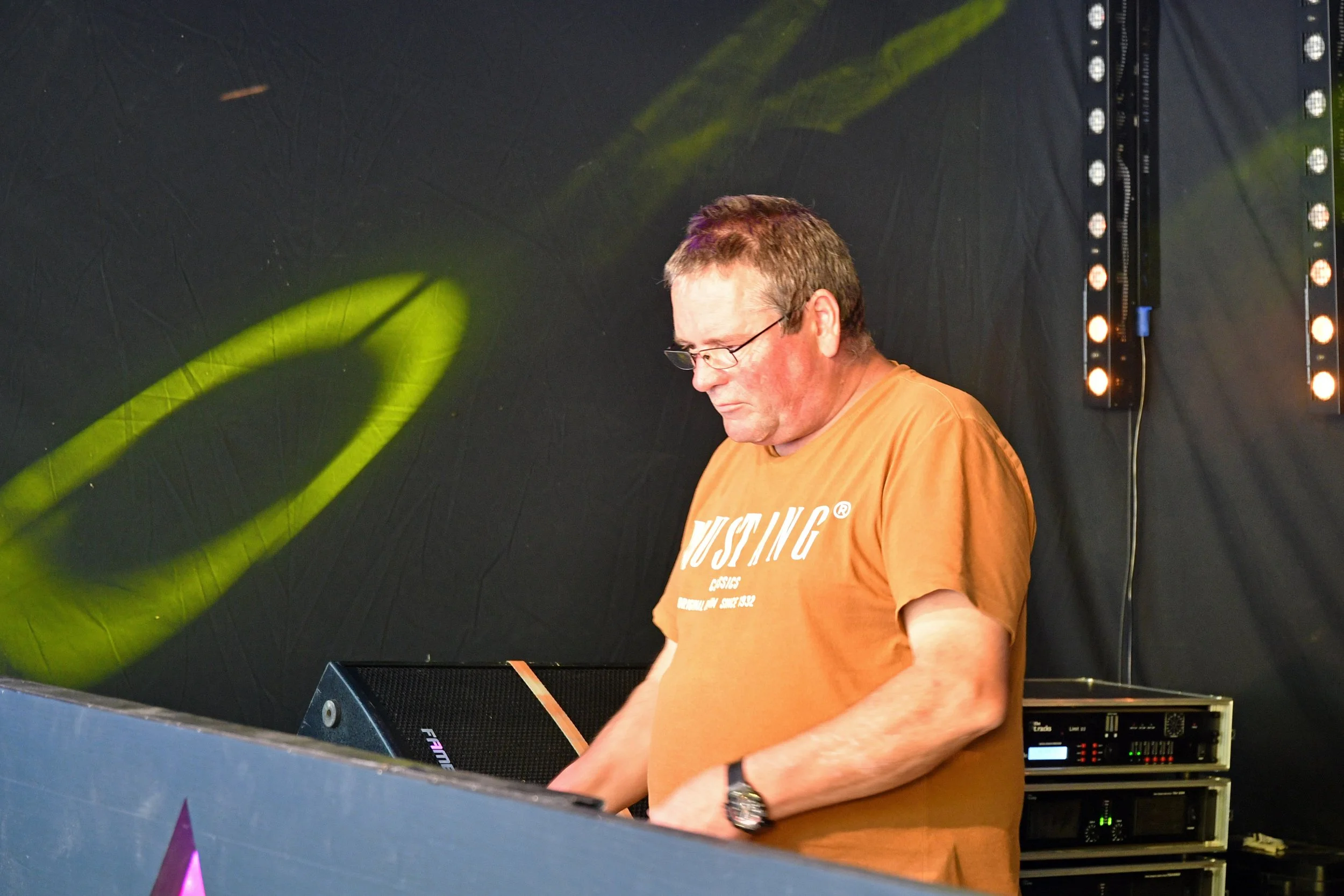 Un homme portant un t-shirt orange avec le mot 'FUSING' est en train de mixer de la musique dans un espace sombre avec un fond noir et un éclairage lumineux.