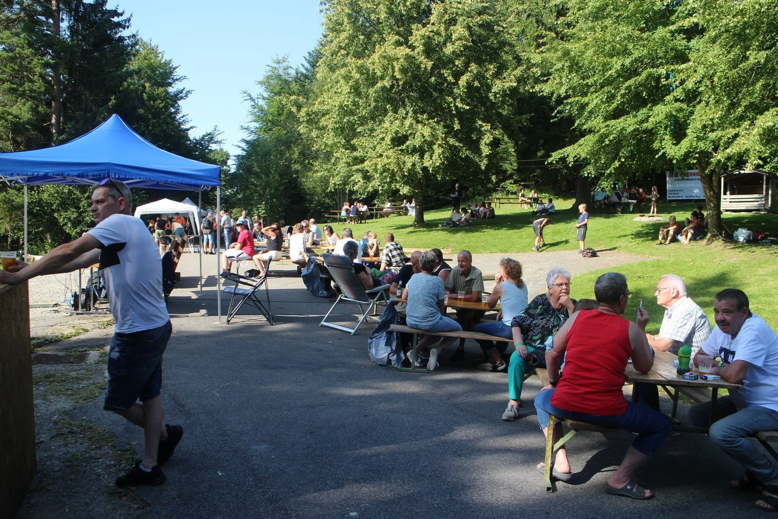 Groupe de personnes assises à des tables en plein air lors d'un événement, avec un espace vert et des arbres en arrière-plan, sous un ciel ensoleillé.