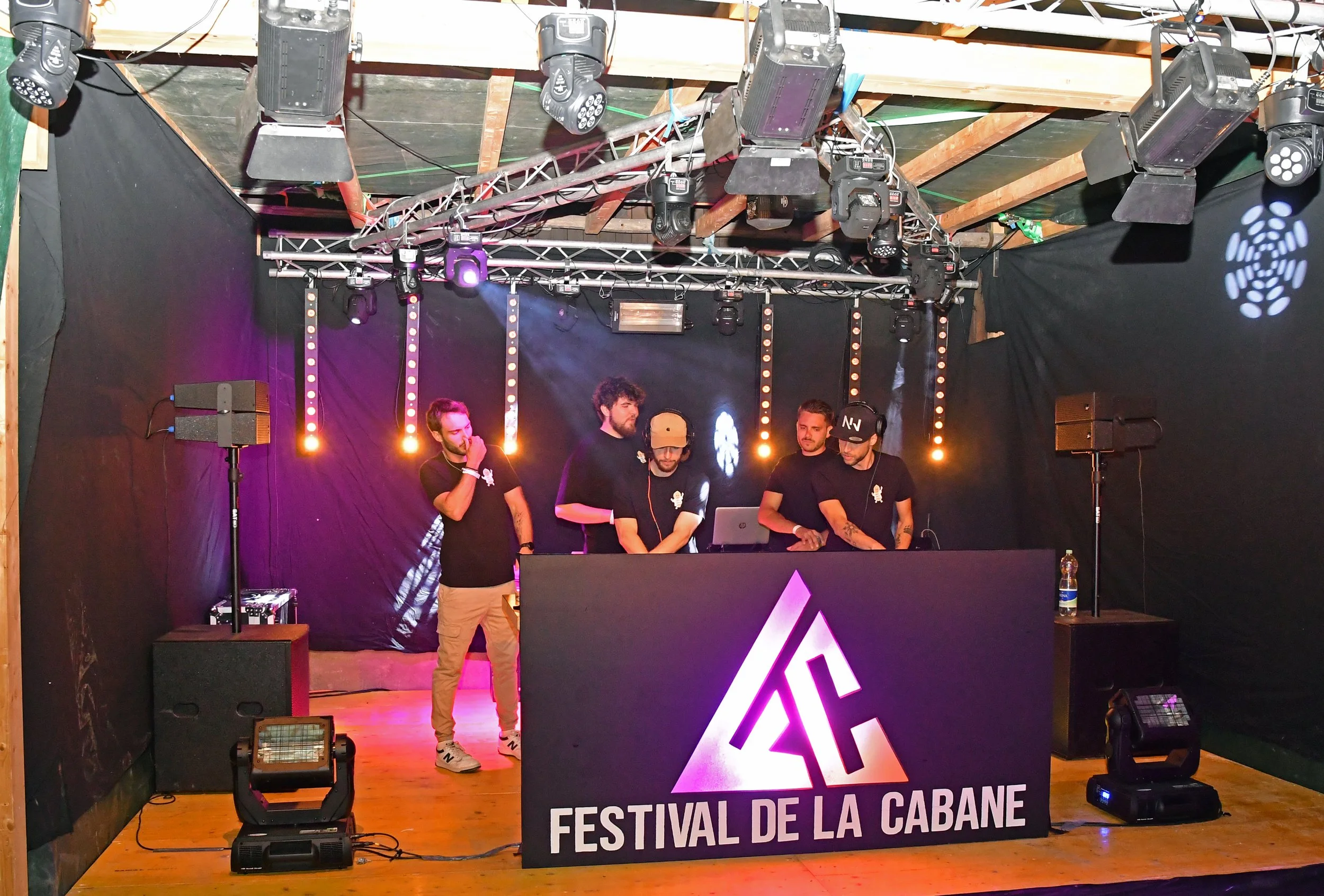 Scène de DJ en train de mixer lors du Festival de la Cabane, avec quatre hommes, équipements de son, éclairage coloré et une bannière avec le nom de l'événement.