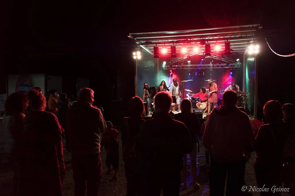 Un groupe de musiciens performe sur une scène éclairée de lumières rouges et multicolores, avec un public regardant la performance lors d’un concert en plein air la nuit.