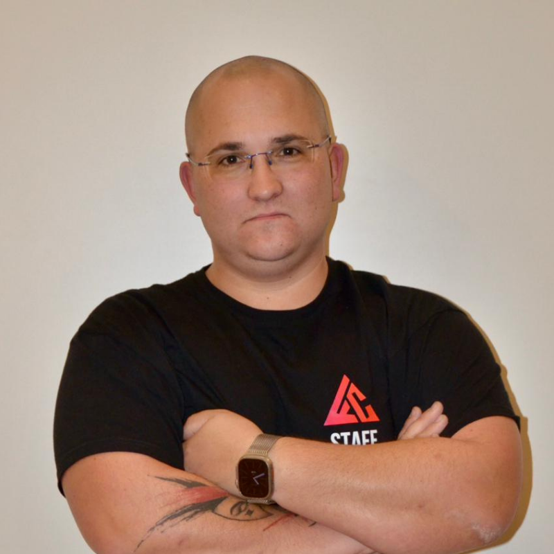 Un homme chauve portant des lunettes, vêtu d'un t-shirt noir avec un logo rouge et blanc, croisant les bras, portant une montre connectée et ayant un tatouage sur le bras gauche.