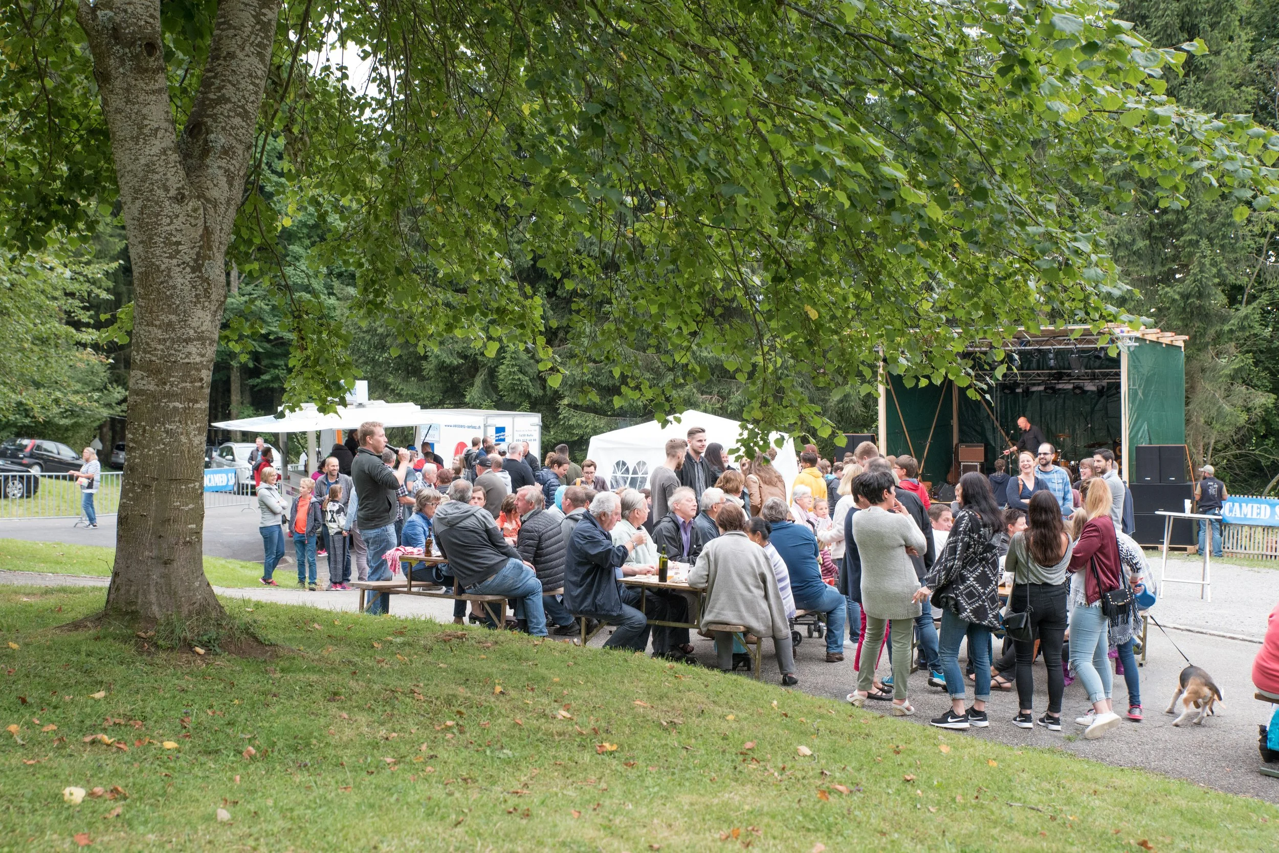Fête en plein air avec un groupe de personnes assises à des tables en bois, un groupe de personnes debout devant une scène en plein air, avec des arbres verts en arrière-plan.