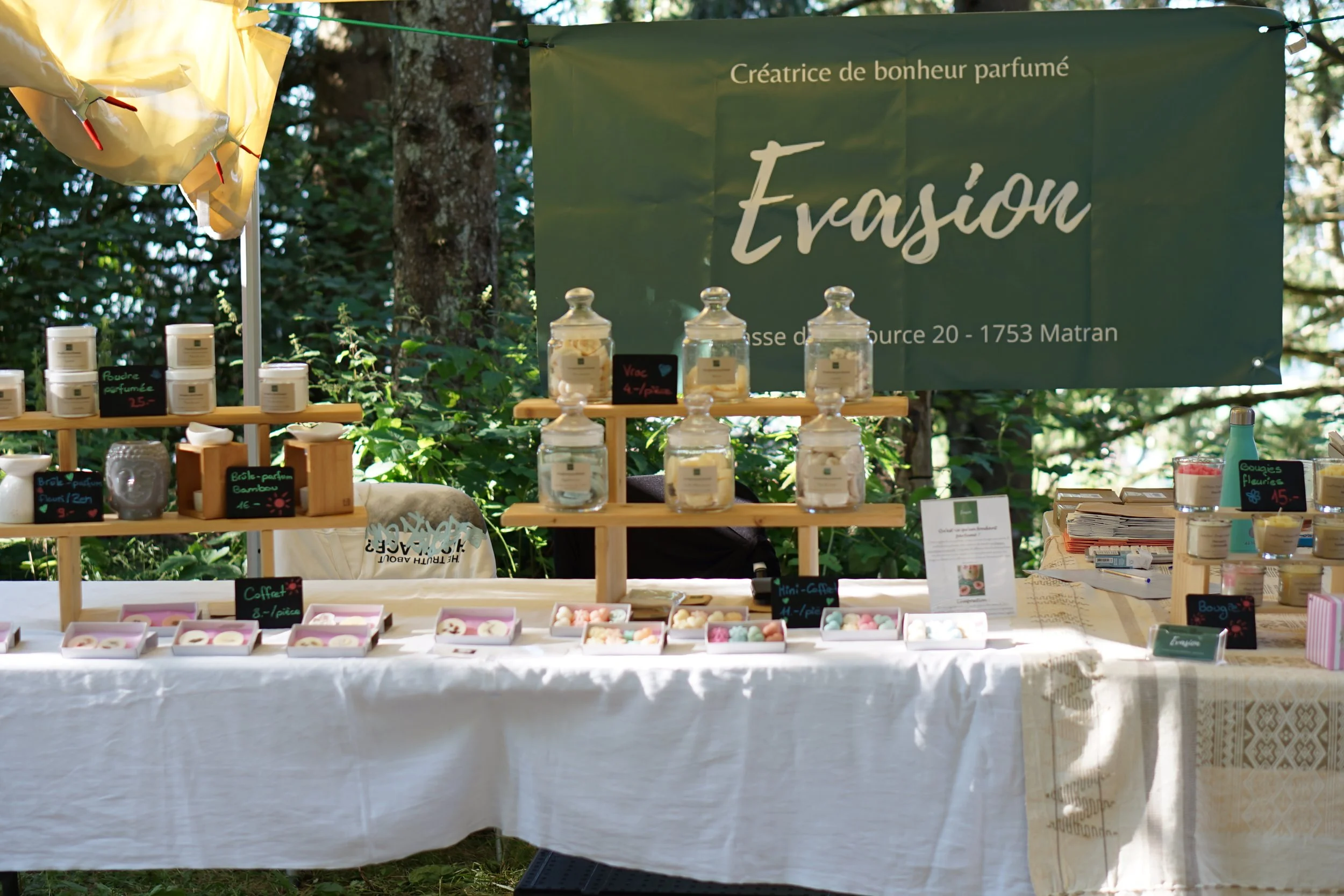 Stand de produits parfumés à l'extérieur avec une bannière verte lisant 'Evasion' et des prix indiqués, entouré d'arbres et de feuillage.