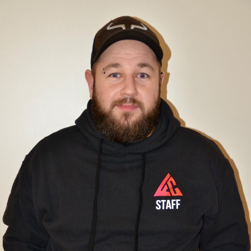 Un homme avec une barbe, portant une casquette et un sweat-shirt noir avec le logo 'STAFF' en rouge et blanc, debout devant un mur blanc.
