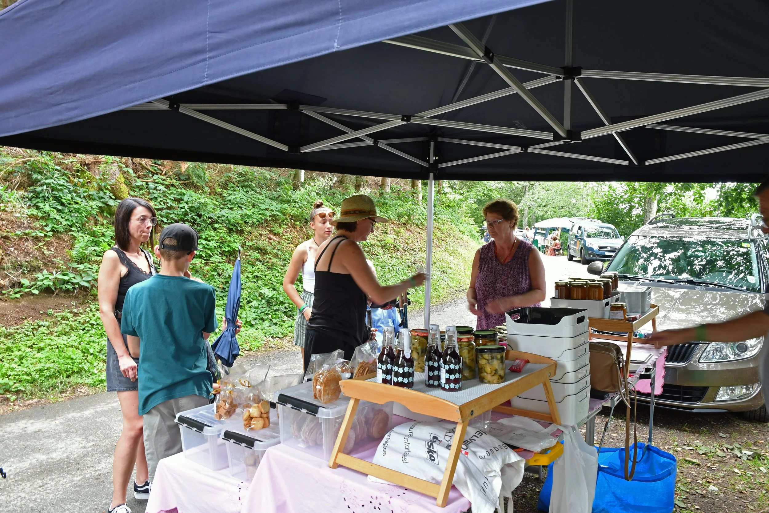 Vendeuse vendant des produits artisanaux sous une tente lors d'un marché en plein air, avec des visiteurs qui attendent.