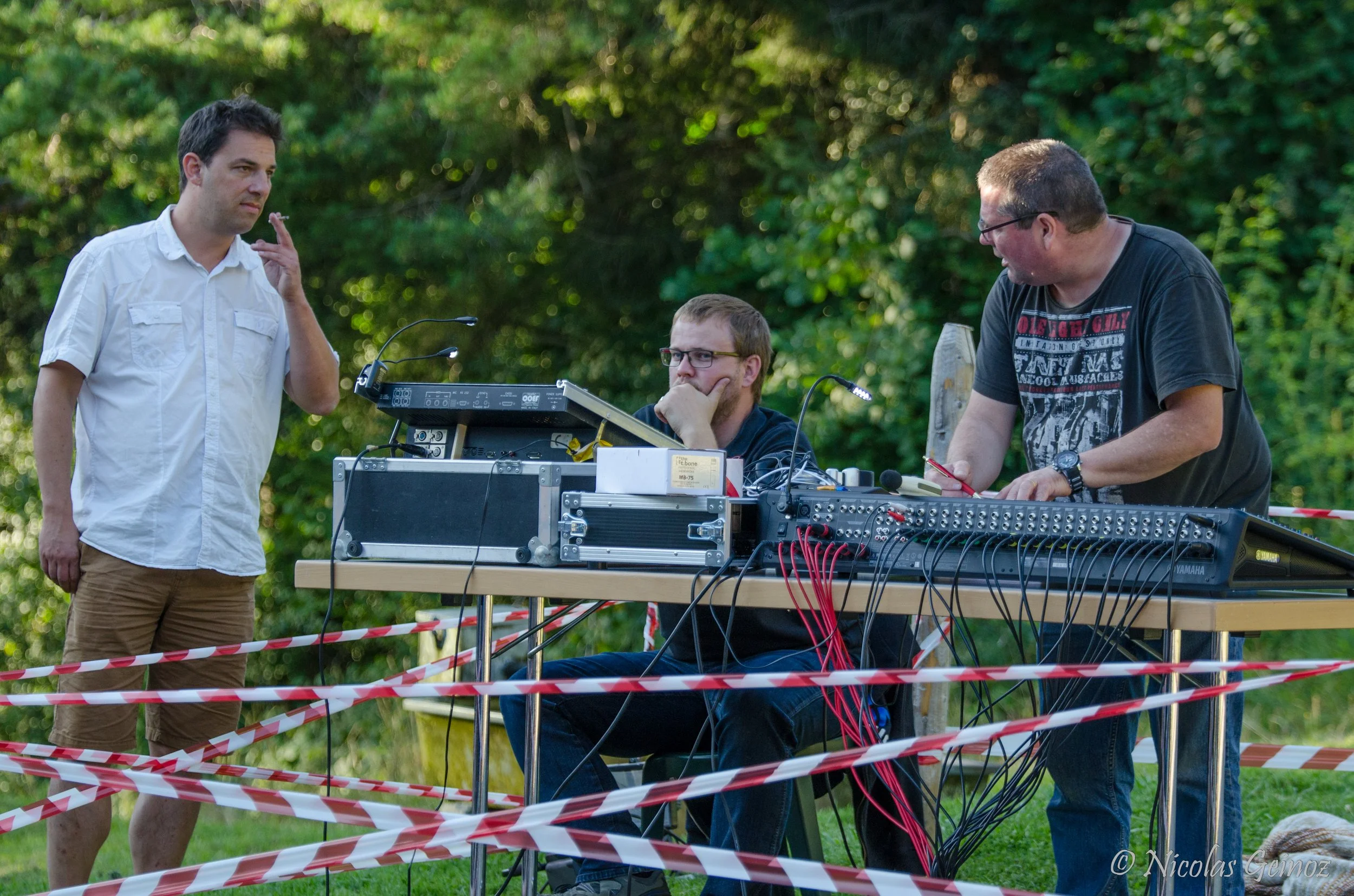 Trois hommes interagissent autour d'une table avec du matériel électronique, en extérieur, avec une zone délimitée par des rubans de signalisation rouges et blancs. Deux hommes se tiennent debout, un à gauche en chemise blanche, l'autre à droite en t