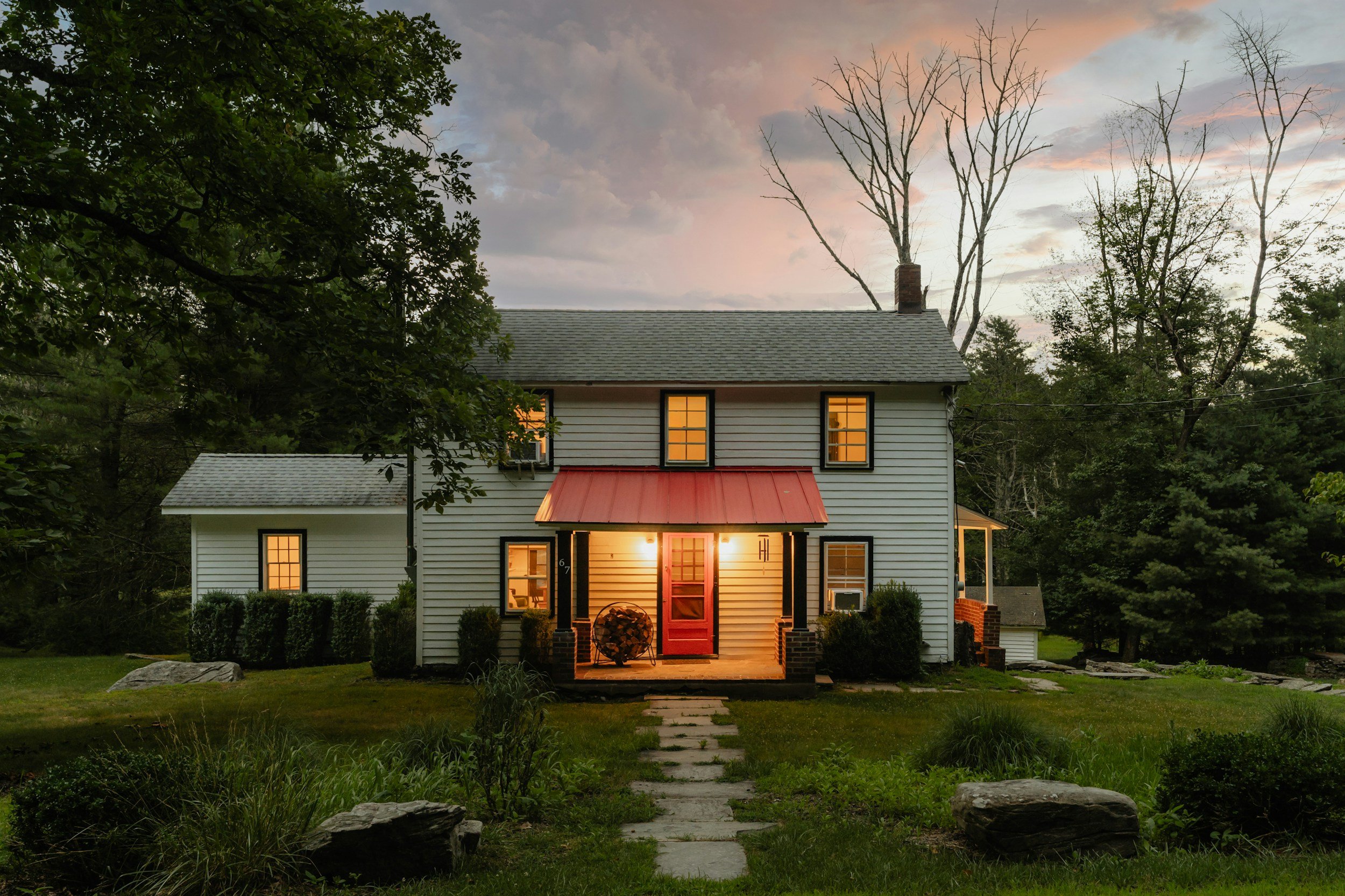 A two-story house with white siding, black window frames, and a red front door illuminated from inside. The house has a small covered porch with black columns and a red roof. The front yard has a stone pathway leading to the porch, surrounded by grass and shrubs, with trees in the background and a colorful sunset sky.