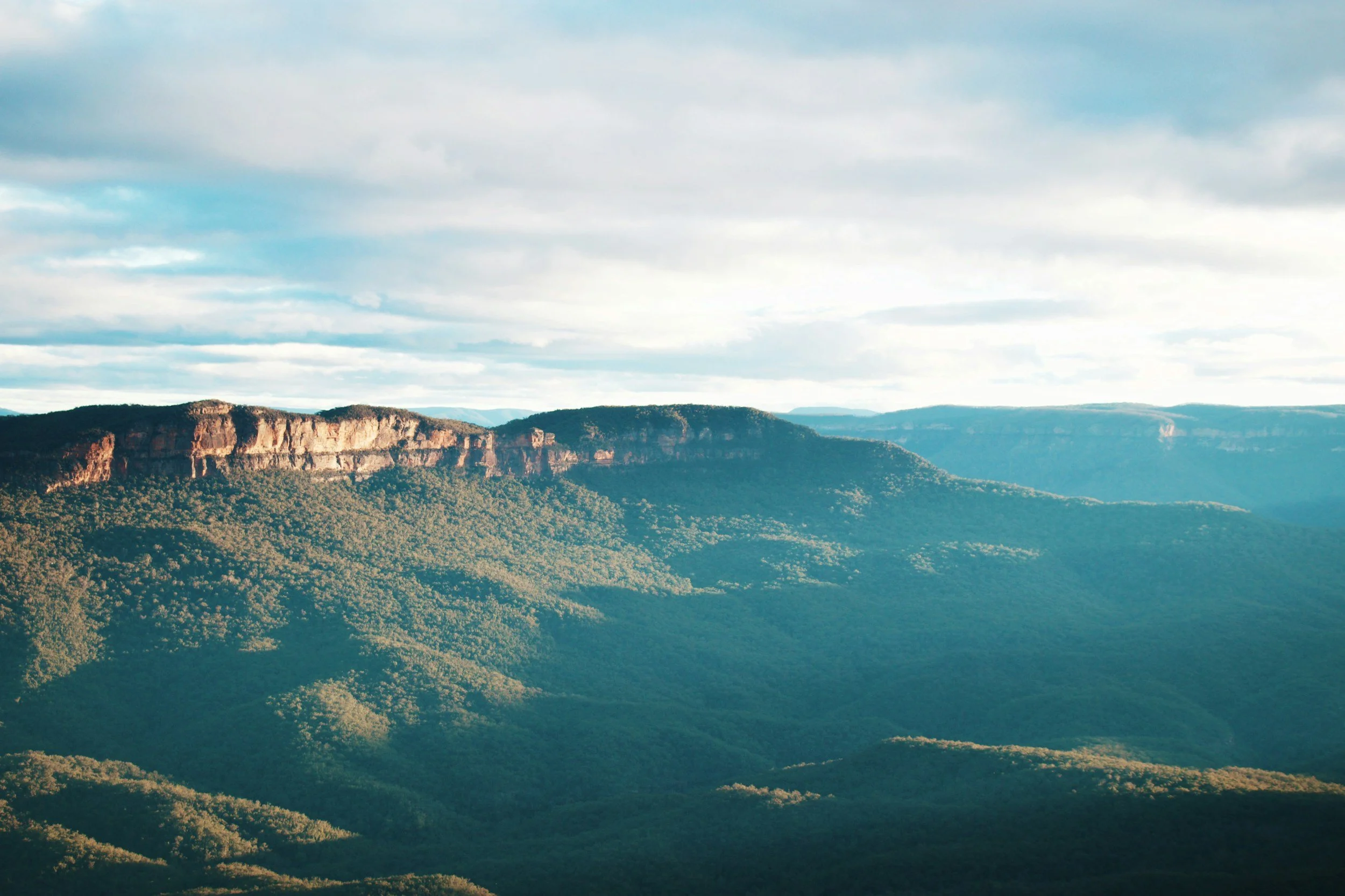 A scenic view of a mountain range with forested slopes and rocky cliffs under a partly cloudy sky.
