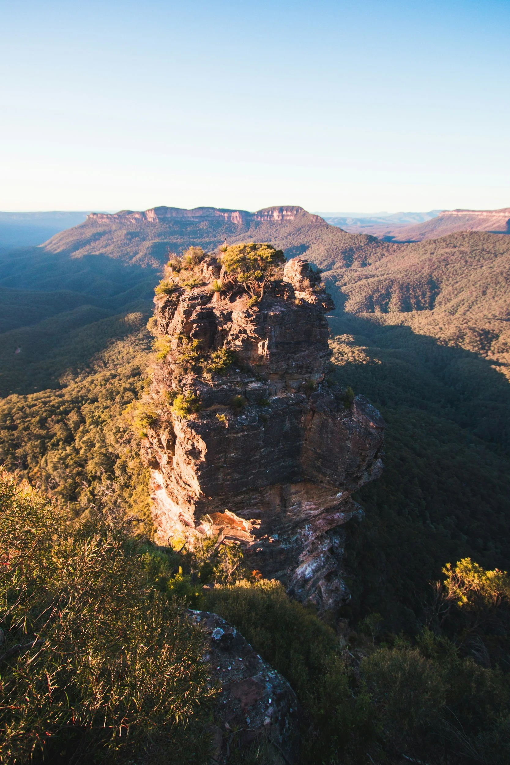 A rugged mountain peak with a large rock formation on top, surrounded by lush green trees and a vast mountain range in the background under a clear blue sky.