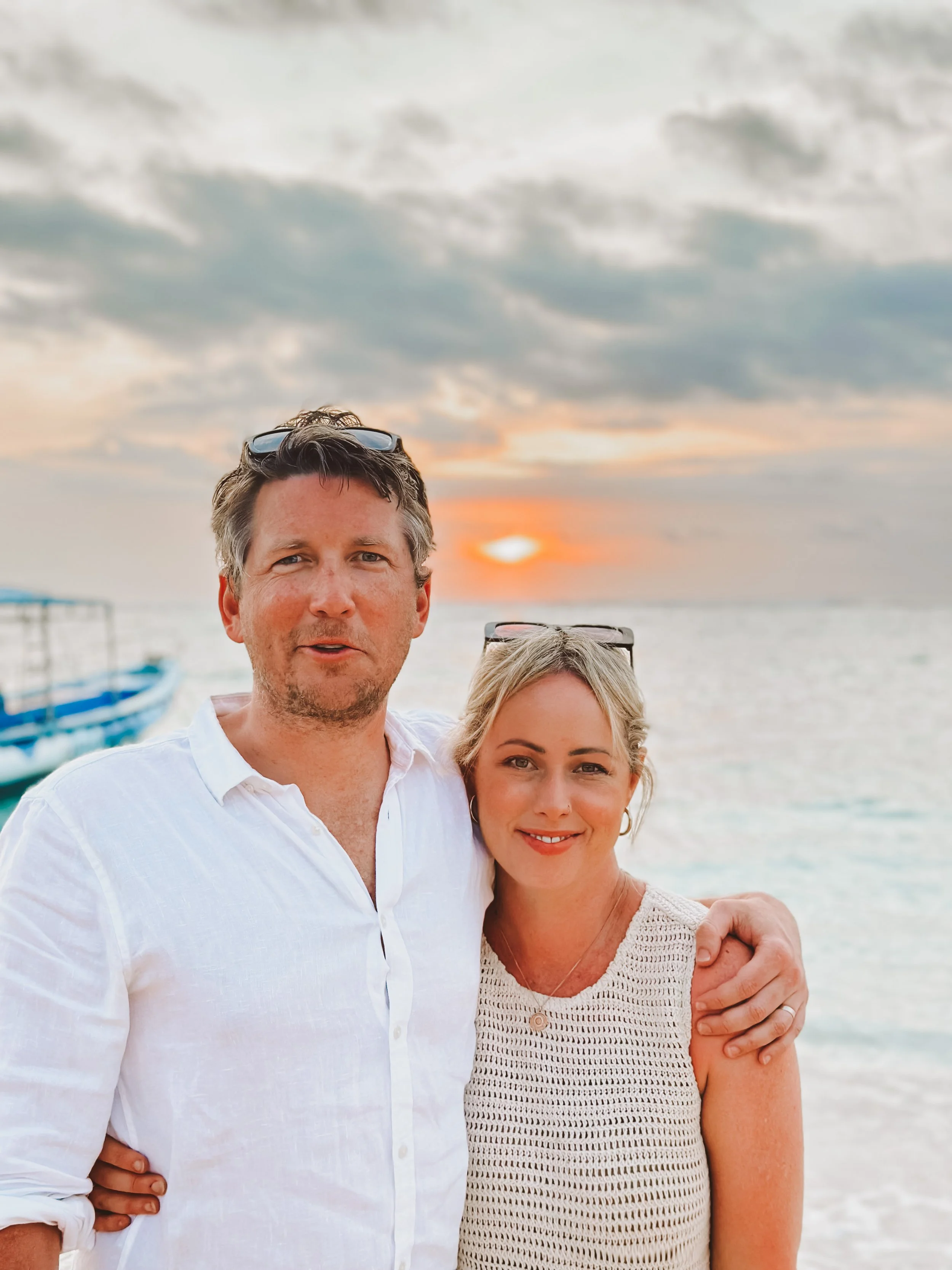 A man and a woman standing close together on a beach at sunset, with the ocean and cloudy sky in the background. They are smiling, with the man's arm around the woman's shoulder.