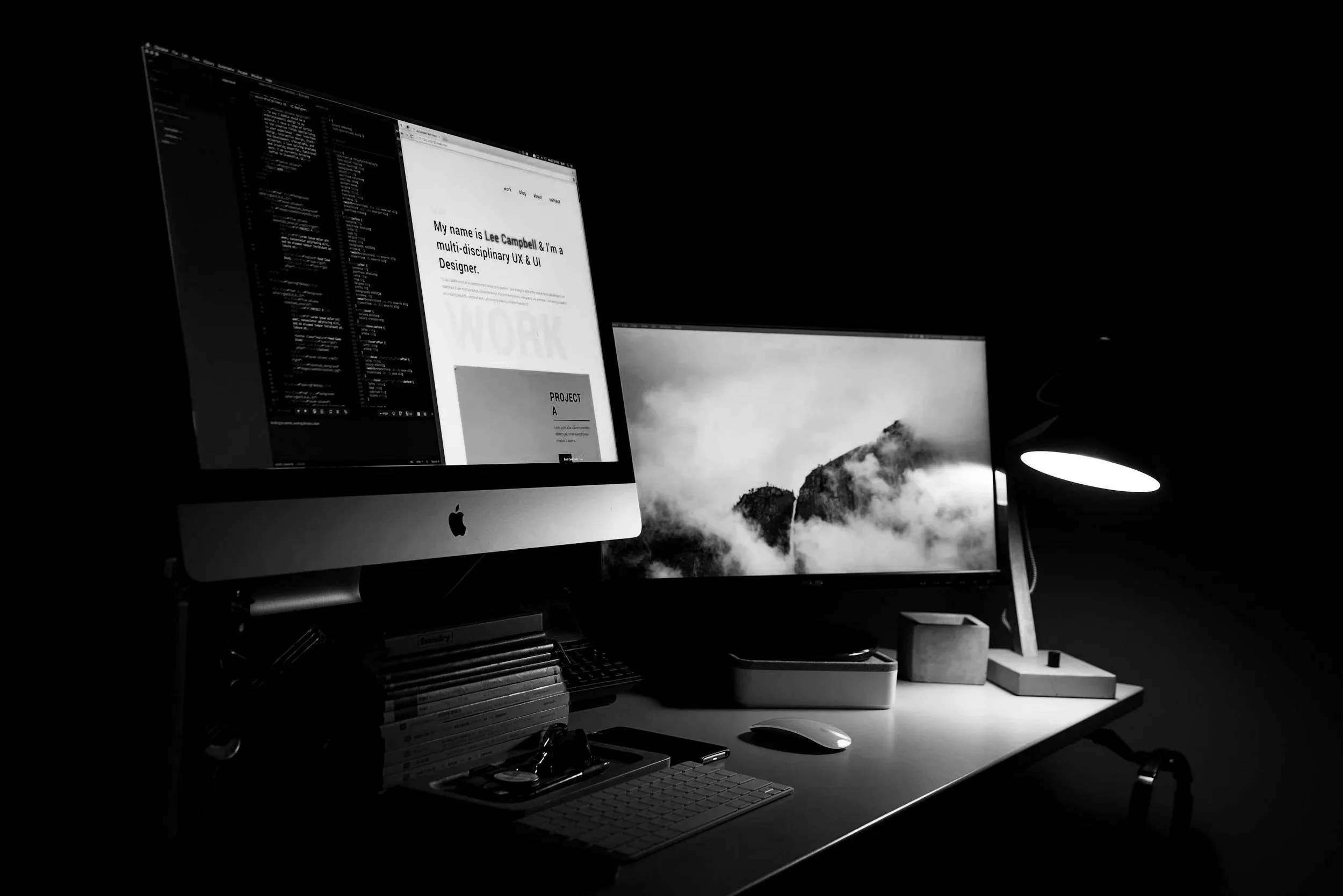 A workspace with an iMac computer displaying a website about UX and UI design, a second monitor showing a mountain scene, a desk lamp, a wireless keyboard, a mouse, and various office supplies in a dimly lit room.