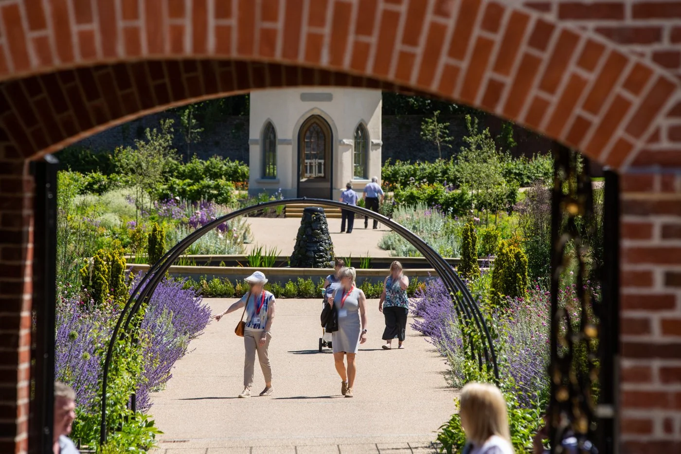 View of a garden with colorful flowers and greenery seen through a brick archway, featuring a small white building in the background, with people walking and enjoying the space on a sunny day.