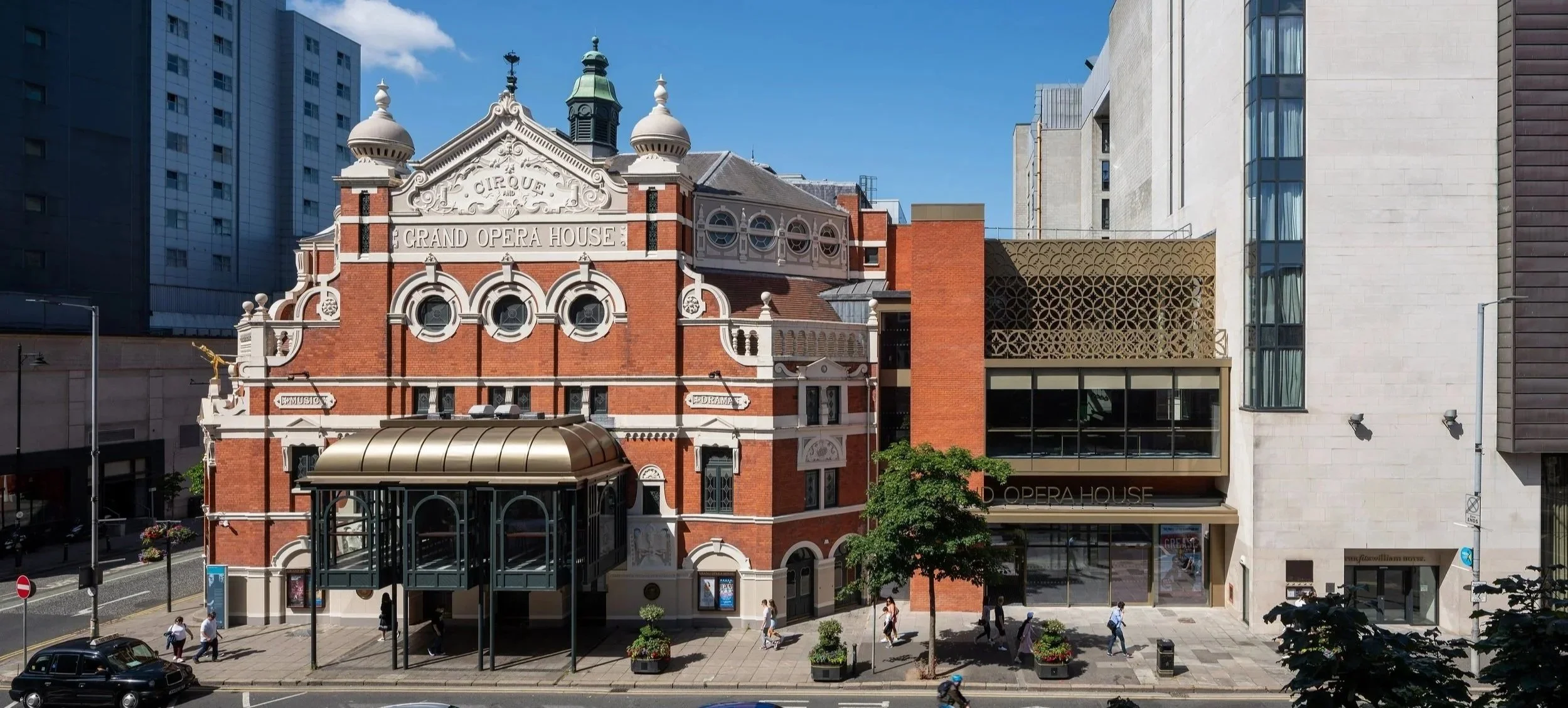 Exterior view of the Grand Opera House building with red brick facade, decorative architectural details, and modern additions, situated on a city street with pedestrians and trees.