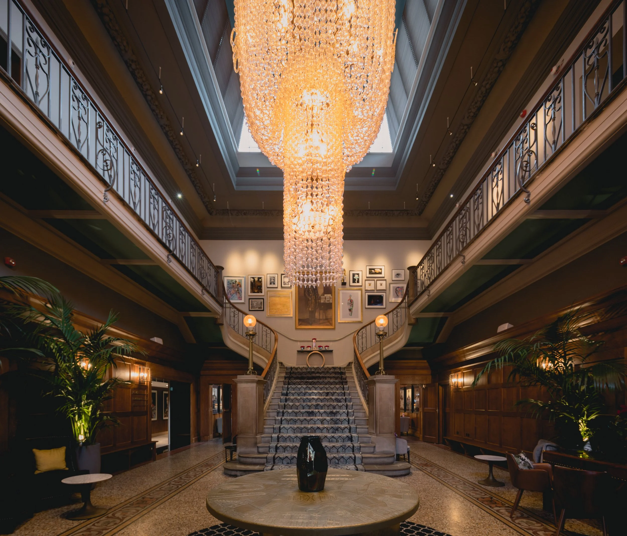Luxurious hotel lobby with grand staircase, ornate chandelier, wall art, and elegant furniture.