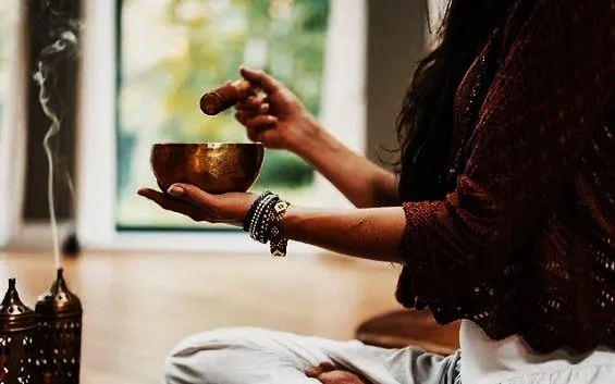 Une personne assise en méditation, tenant une cloche tibétaine dans ses mains, près d'une fenêtre avec une vue extérieure verdoyante.