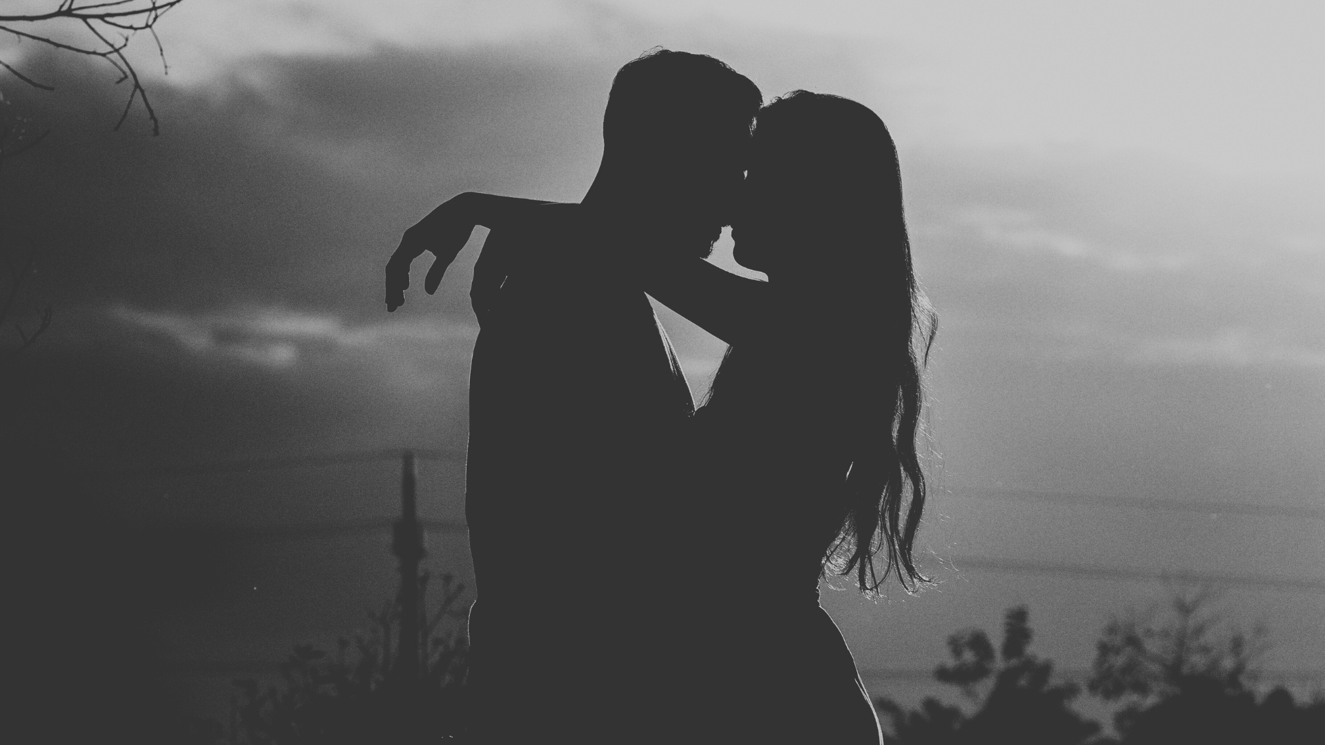 Silhouette of a couple embracing and touching foreheads outdoors at sunset, with trees and power lines in the background.