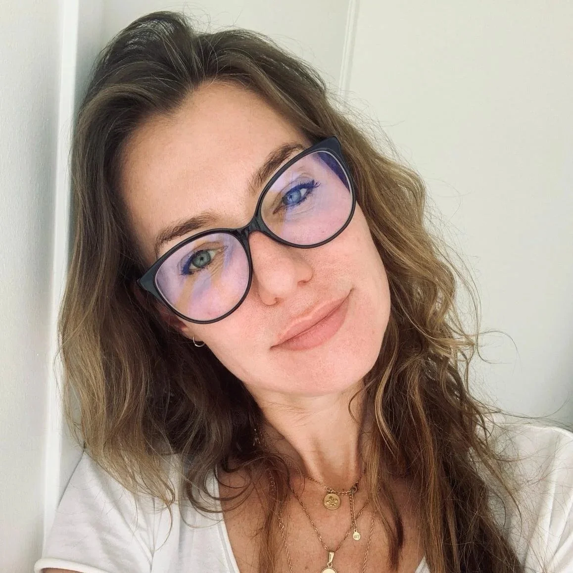Close-up of a woman with wavy brown hair, wearing large black-rimmed glasses, multiple gold necklaces, and a white top, leaning against a light-colored wall, smiling gently.