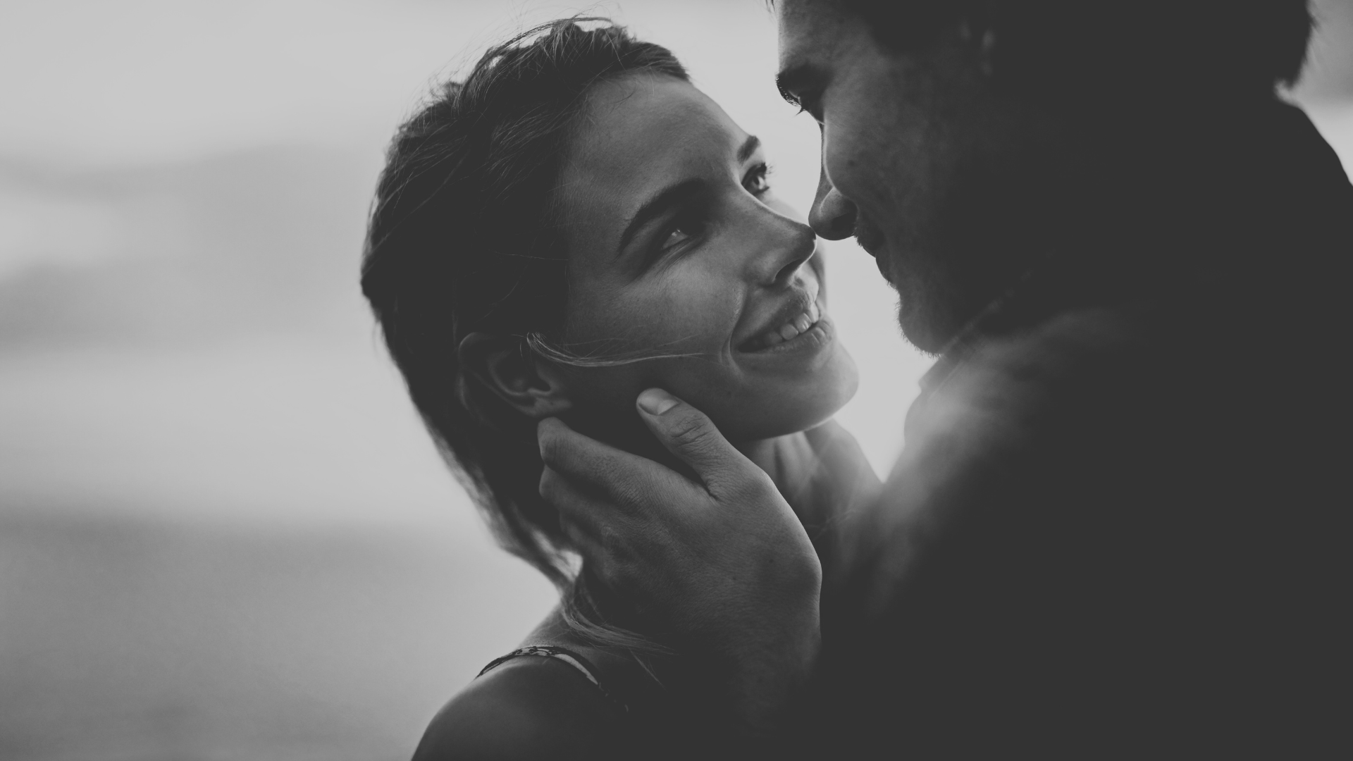 A black and white photo of a couple close together, smiling and looking into each other's eyes, with the woman gently holding the man's face.