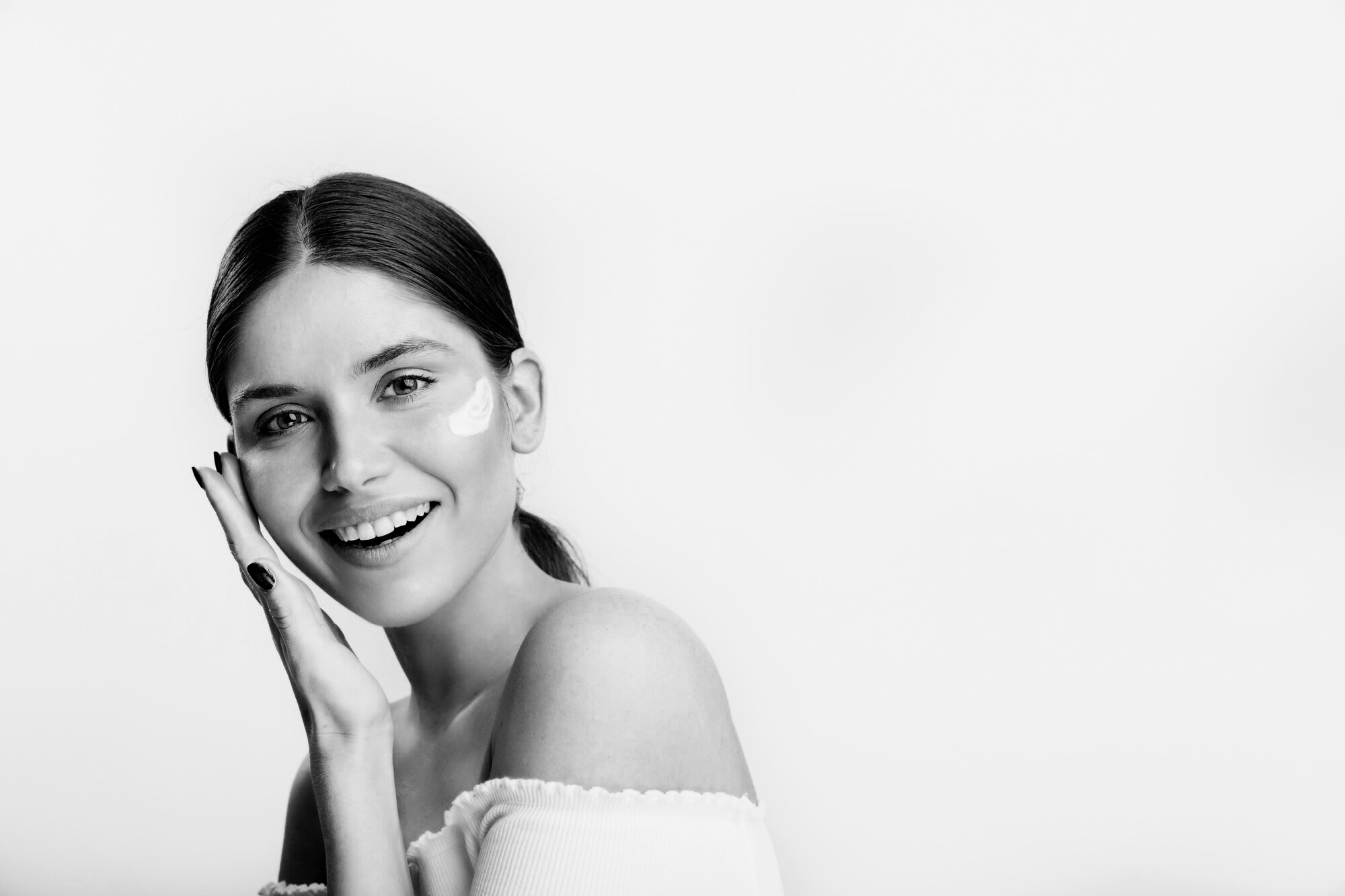 Ragazza con capelli raccolti, che sorride e si tocca il viso con la mano, senza trucco visibile, in bianco e nero.