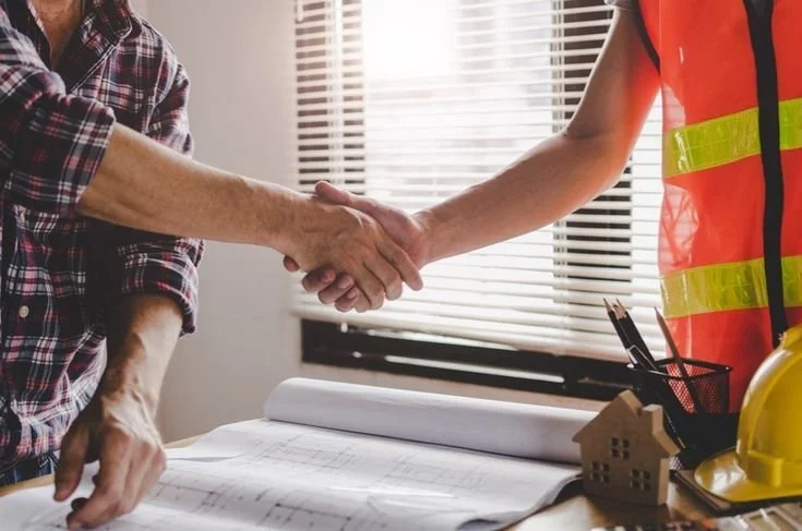 Two people shaking hands over a table with blueprints, a small house model, and a yellow safety helmet, with window blinds in the background.