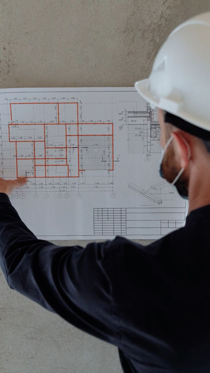 A person wearing a safety helmet and mask pointing at a technical construction blueprint with multiple rooms outlined in red.