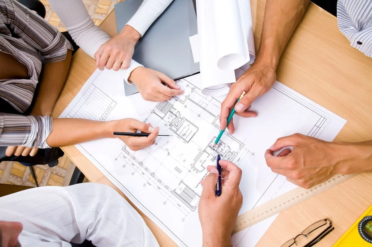 People working on architectural blueprints at a desk with rulers, glasses, and rolled-up papers.