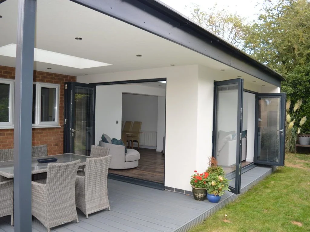 A modern house with a white exterior, outdoor patio with wicker chairs and a dining table, and large glass doors that open to a living room inside. There are a few potted plants outside.