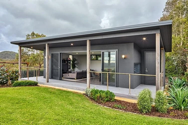 Modern house with gray exterior, large open patio with seating, surrounded by green lawn and bushes, trees in the background, overcast sky.
