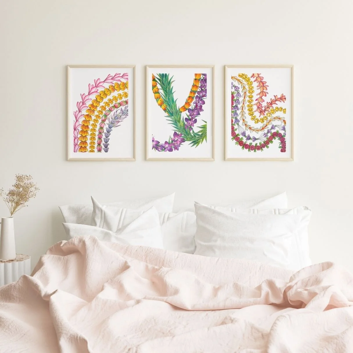 White wall with three framed floral Lei prints, over a bed and a small side table with a dried flower arrangement.