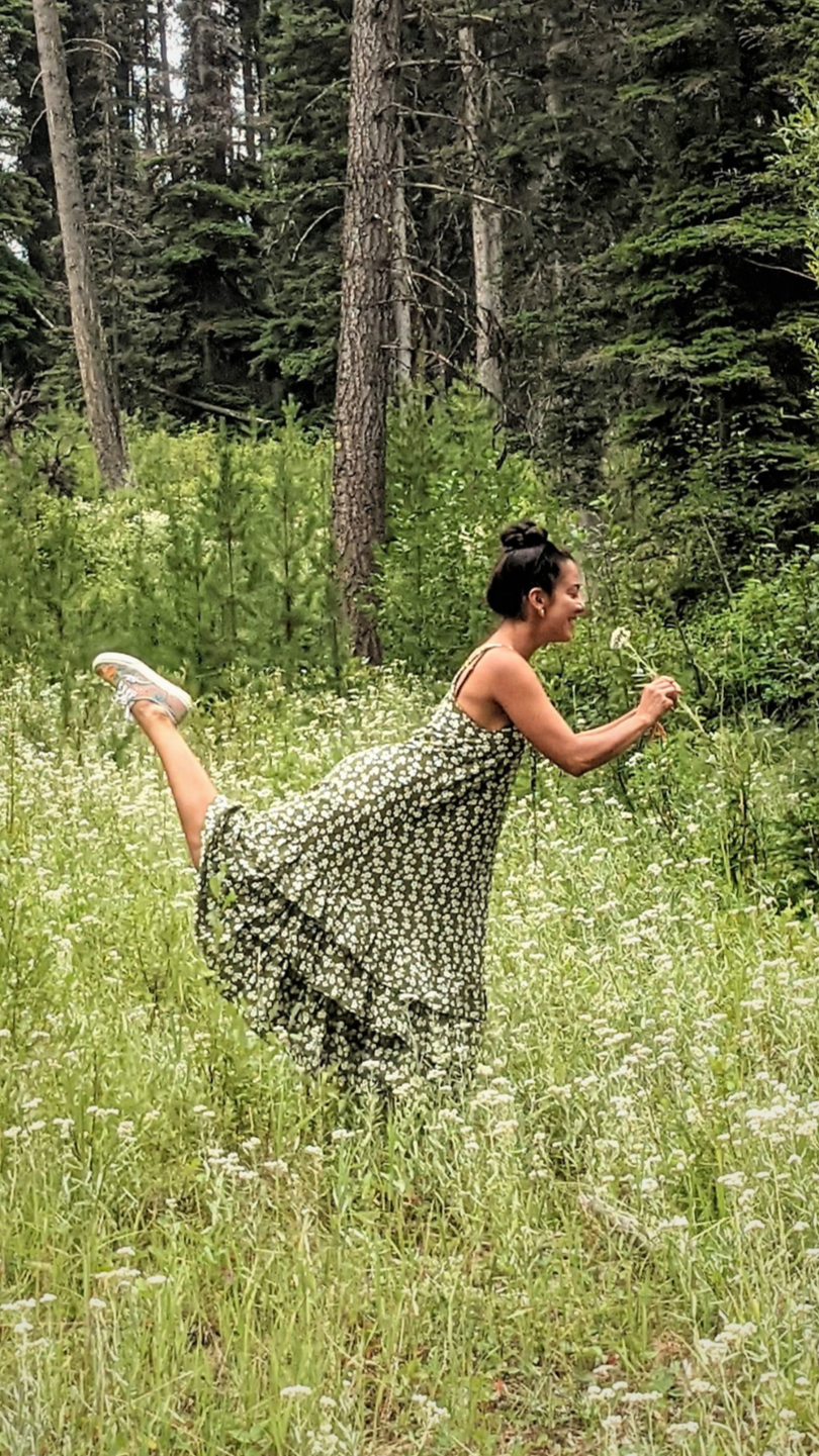 Artist in a floral dress is in a forested area with tall trees and green foliage, holding a flower and smiling while posing through a grassy meadow of small white flowers.