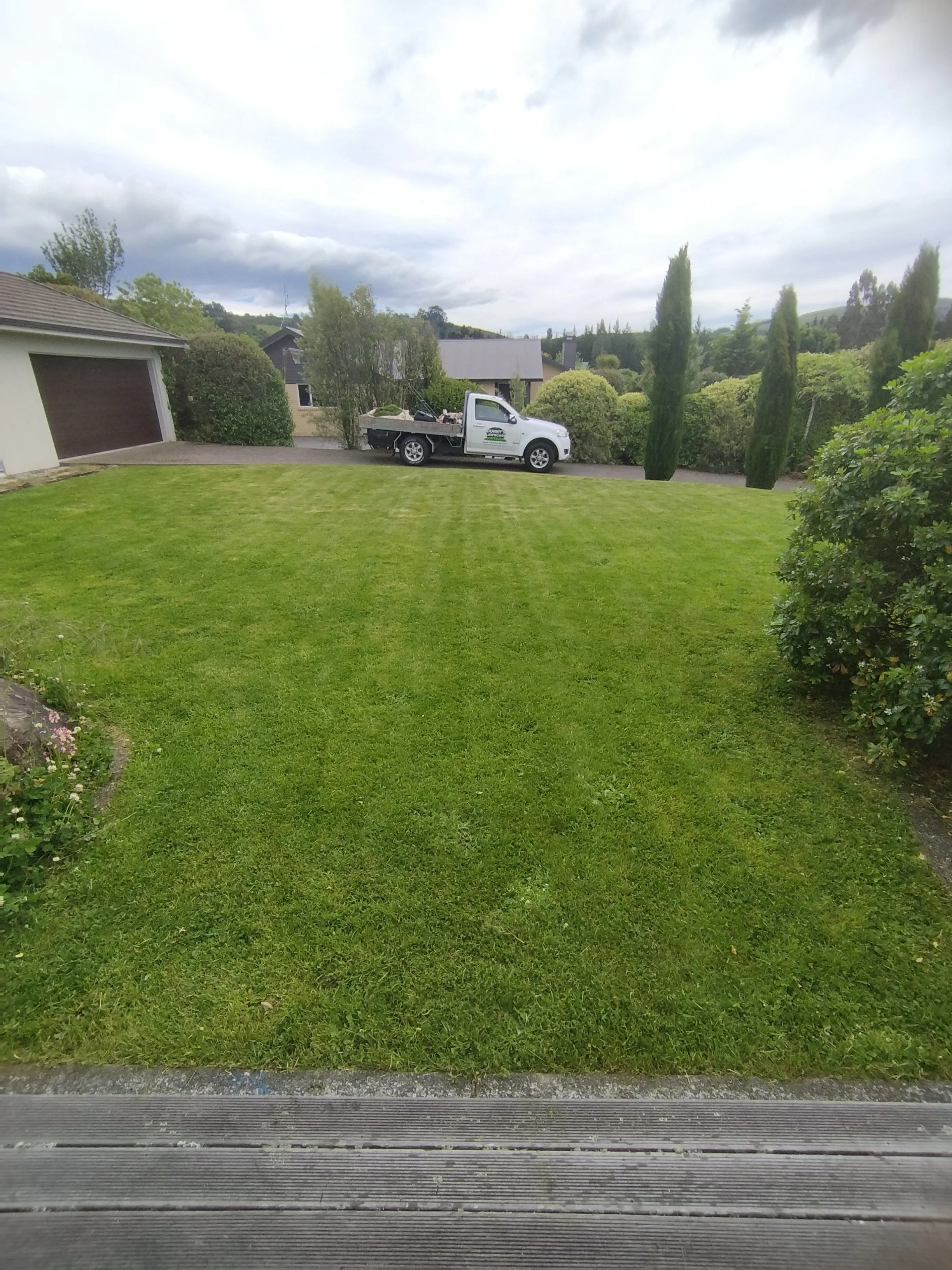 View of a well-maintained green lawn with a driveway at the back, featuring a white pickup truck parked along it, surrounded by trees and bushes, under a cloudy sky.
