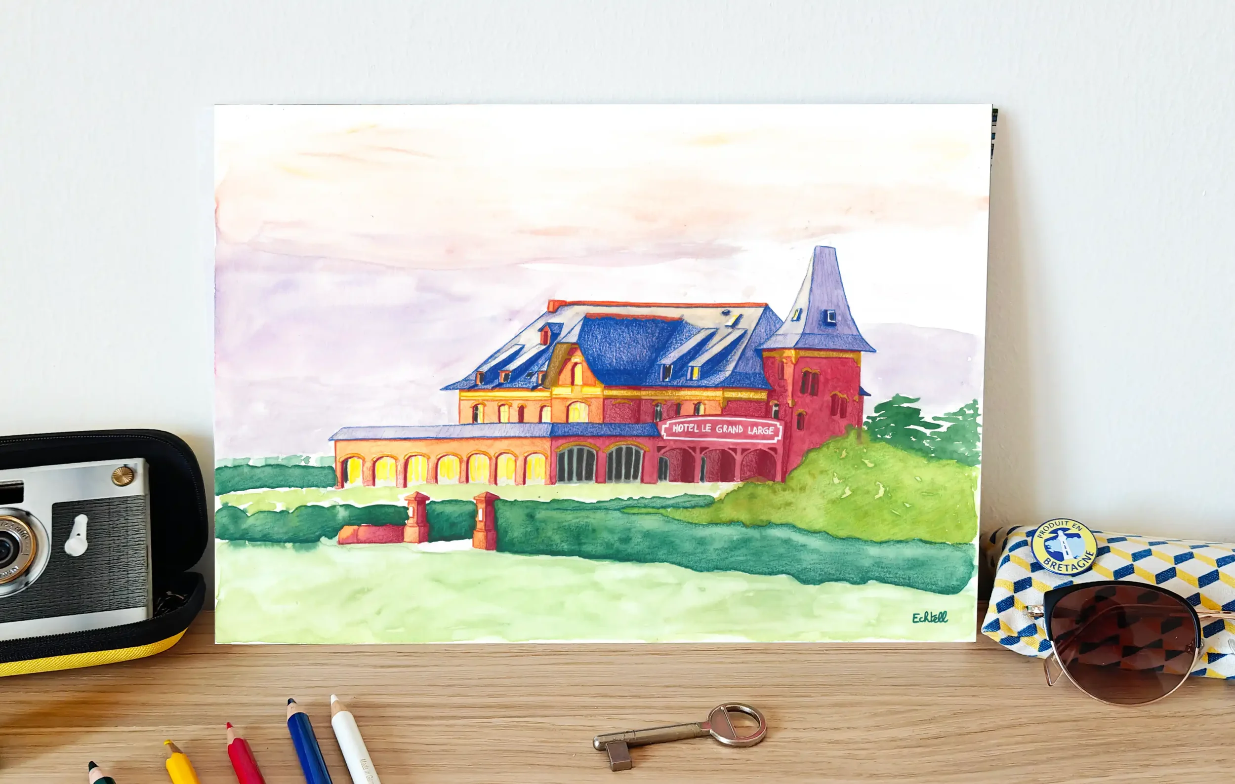 Affiche de l'hôtel Le Grand Large posée sur un meuble en décoration murale avec des lunettes de soleil, crayons de couleur, clé, photo. Dessin à la main hôtel de bord de mer côte atlantique Bretagne Loire Atlantique tourisme. Echtell Art illustration