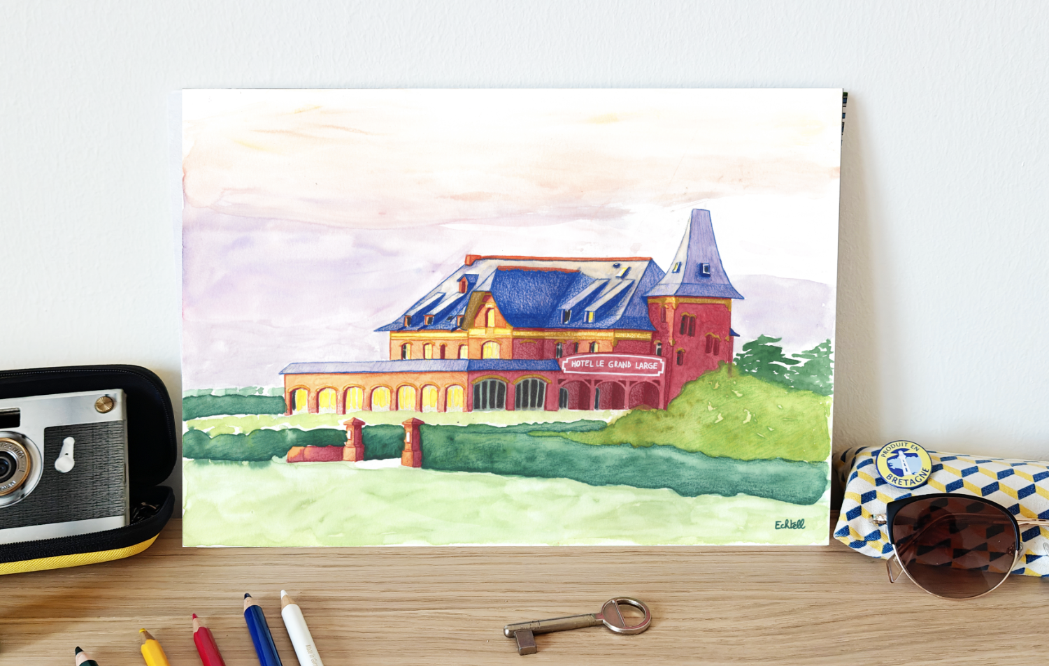 Affiche de l'hôtel Le Grand Large posée sur un meuble en décoration murale avec des lunettes de soleil, crayons de couleur, clé, photo. Dessin à la main hôtel de bord de mer côte atlantique Bretagne Loire Atlantique tourisme. Echtell Art illustration
