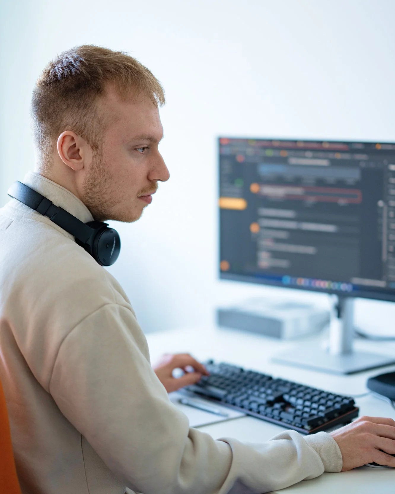 Junger Mann arbeitet am Computer mit Monitor, Keyboard und Maus, trägt Kopfhörer um den Hals, im Hintergrund ein heller Raum.