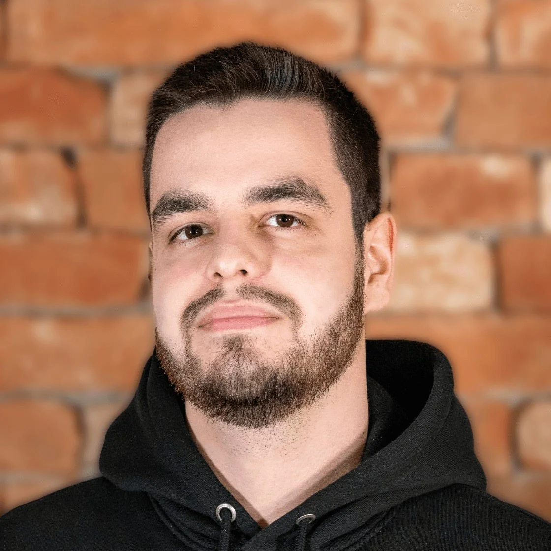 Porträt eines Mannes mit dunklem Haar, Bart, graublauen Augen, trägt schwarzen Kapuzenpullover, vor einer Ziegelsteinwand.