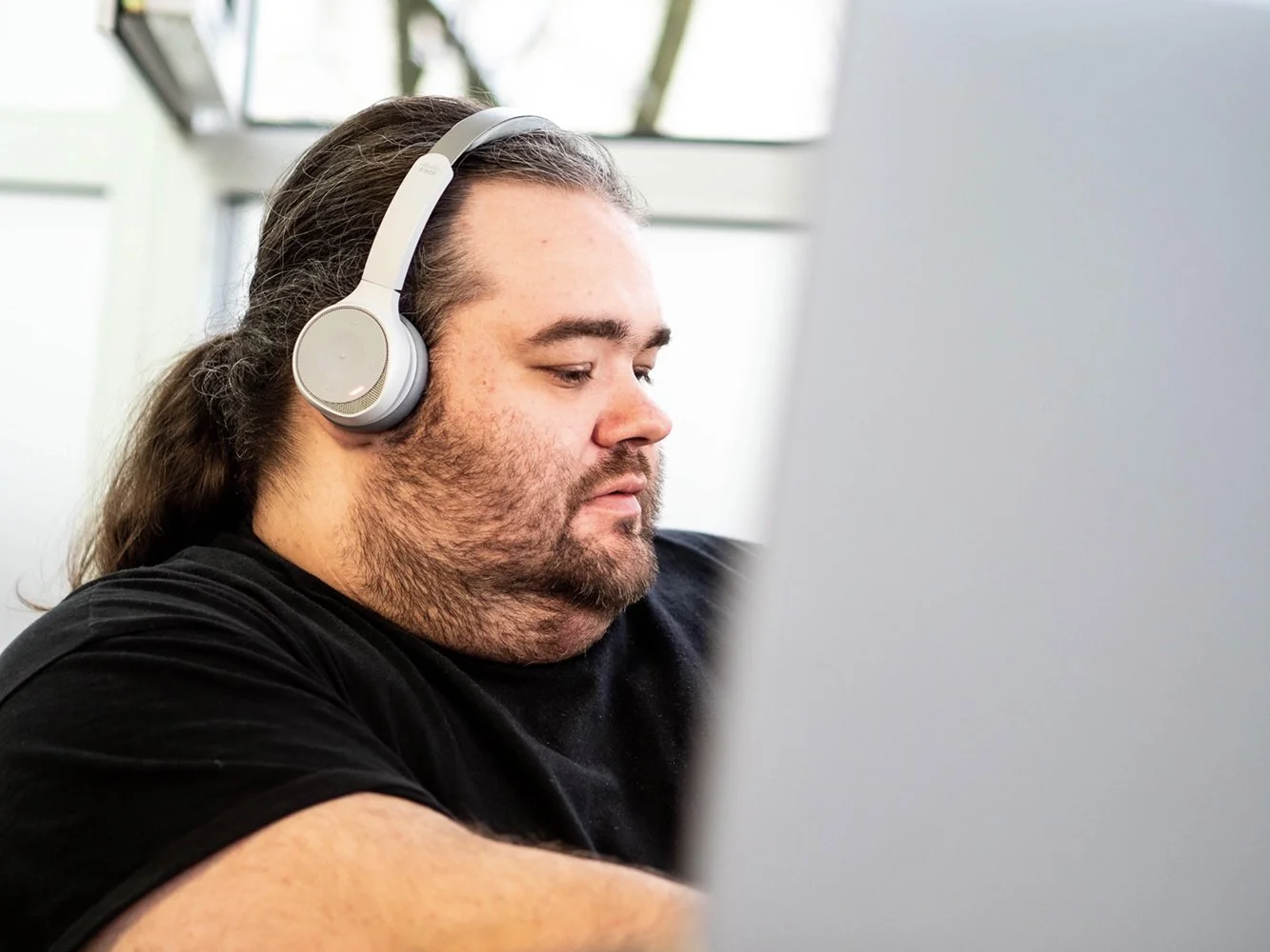 Ein Mann mit langen Haaren und Bart trägt Kopfhörer und arbeitet an einem Computer in einem hellen Raum, wahrscheinlich ein Büro.