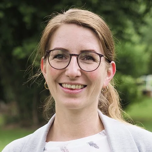 Ein Lächeln einer Frau mit Brille vor einem grünen, naturbelassenen Hintergrund.