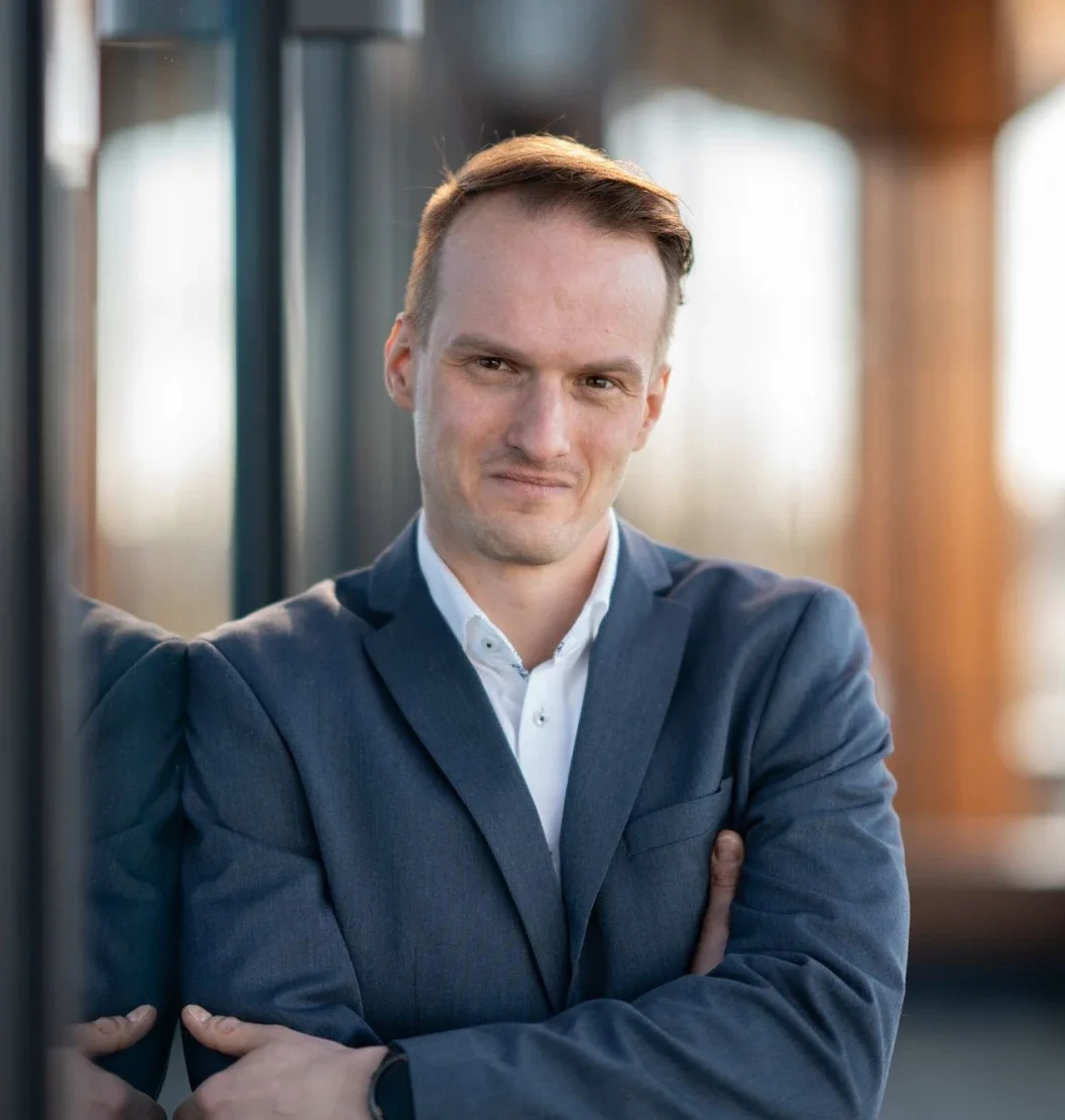 Ein Mann in einem dunklen Anzug mit weißen Hemd, der selbstbewusst mit verschränkten Armen in einem modernen Büro steht.